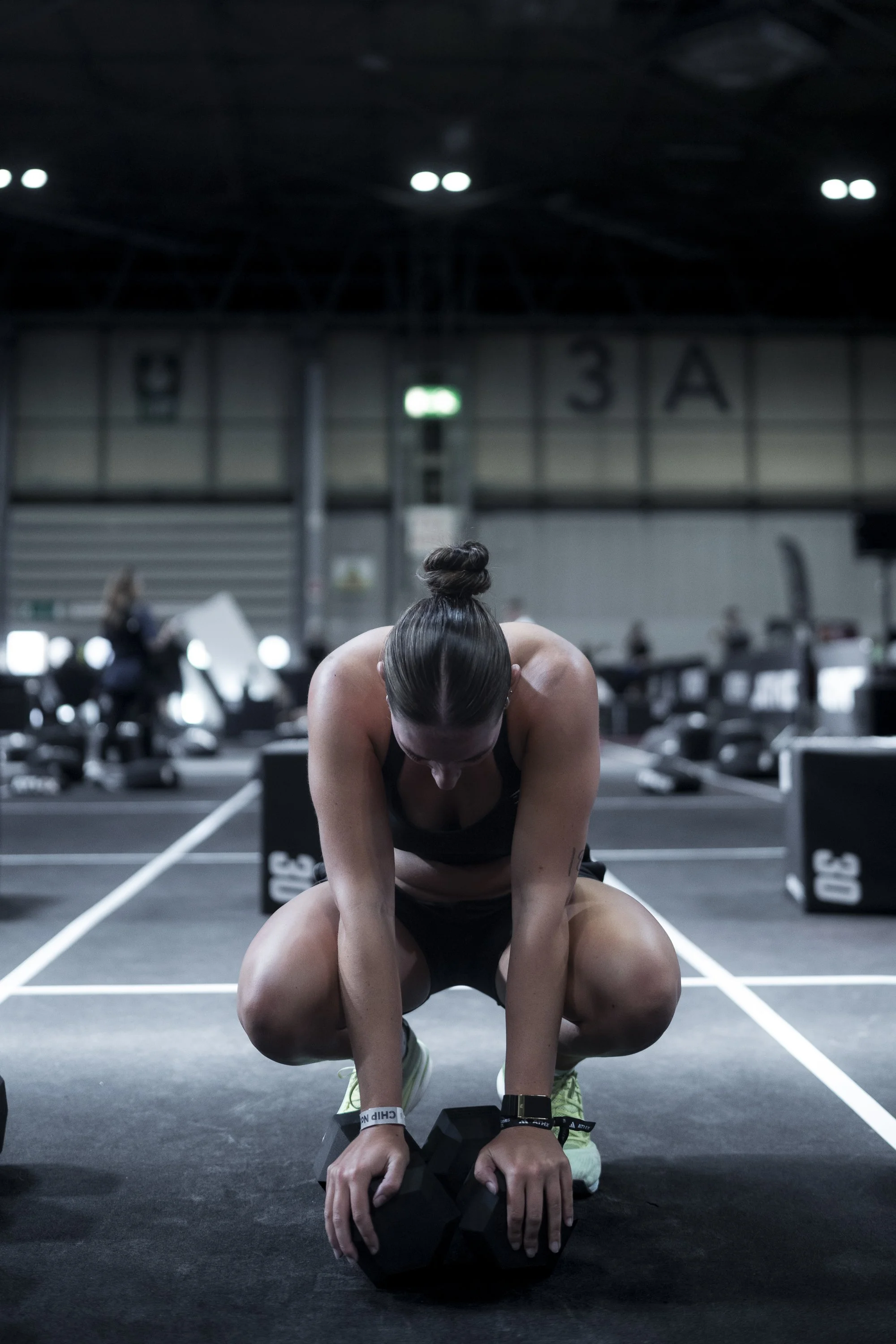 A woman is kneeling on a gym floor, leaning forward with her hands on a large black weight, appearing to be preparing for a workout, with gym equipment and other people in the background.