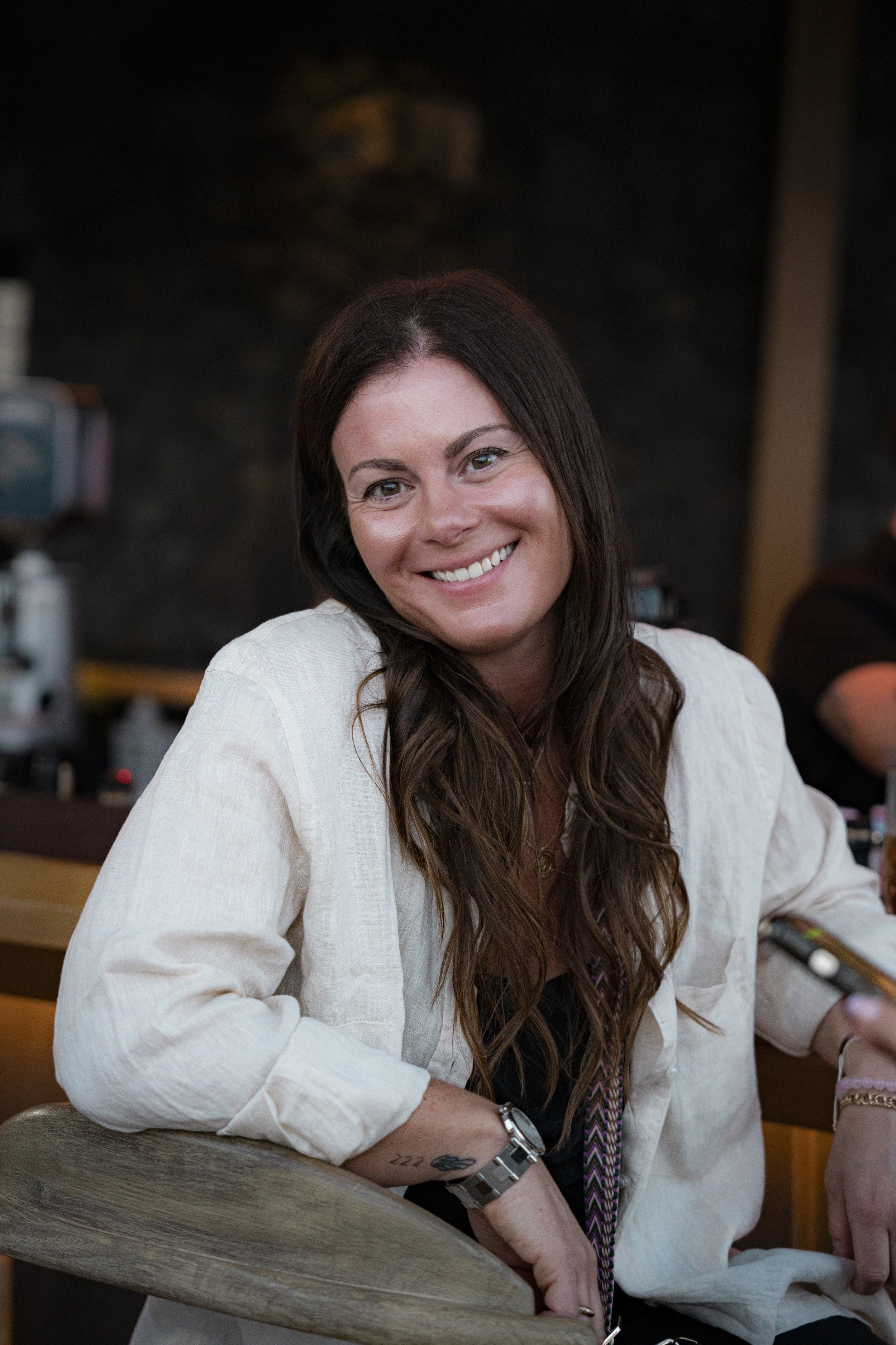 A woman with long brown hair smiling at the camera, sitting at a bar or restaurant with a dark background.