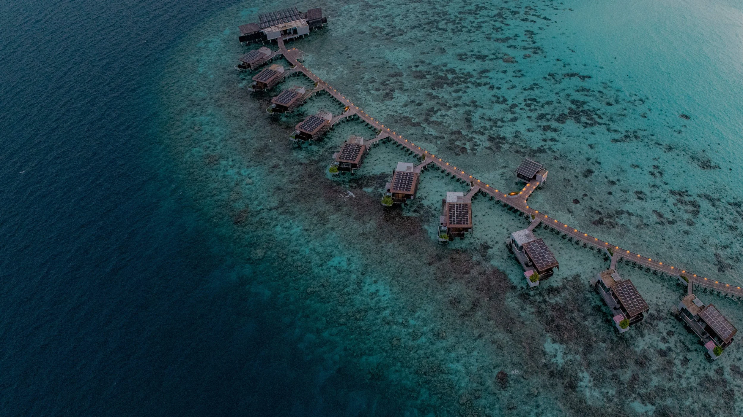 Overwater bungalows with solar panels, connected by a walkway, at a tropical resort over a coral reef.