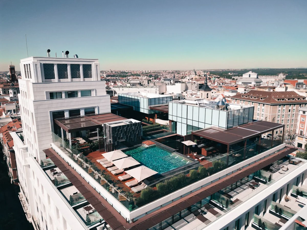 Aerial view of a modern hotel rooftop with a swimming pool, sun loungers, umbrellas, and glass-walled structures, city skyline in the background.