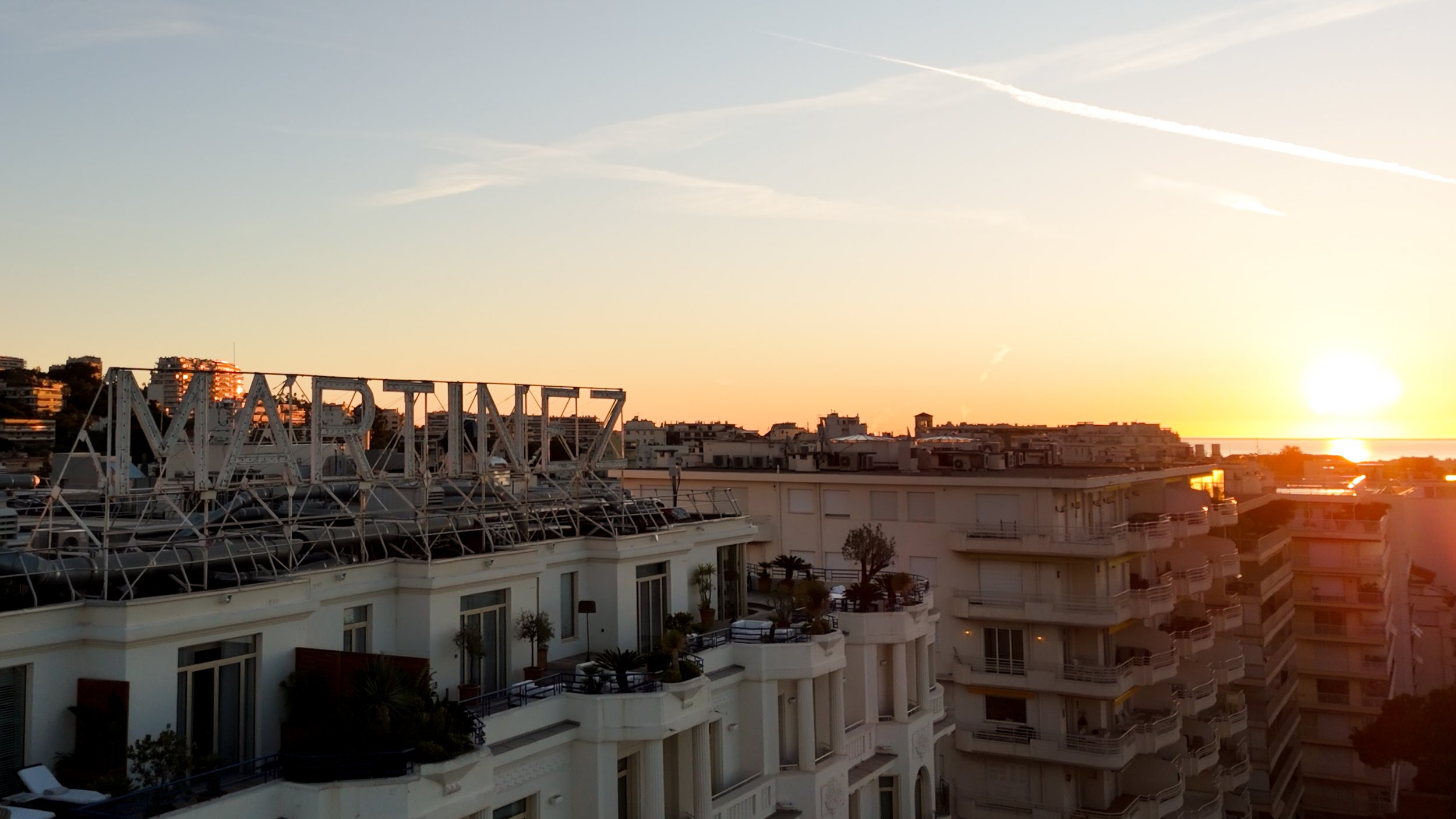 Sunset over a cityscape with white modern buildings, some balconies with plants, and a large rooftop sign that reads 'MARINA' in the foreground.