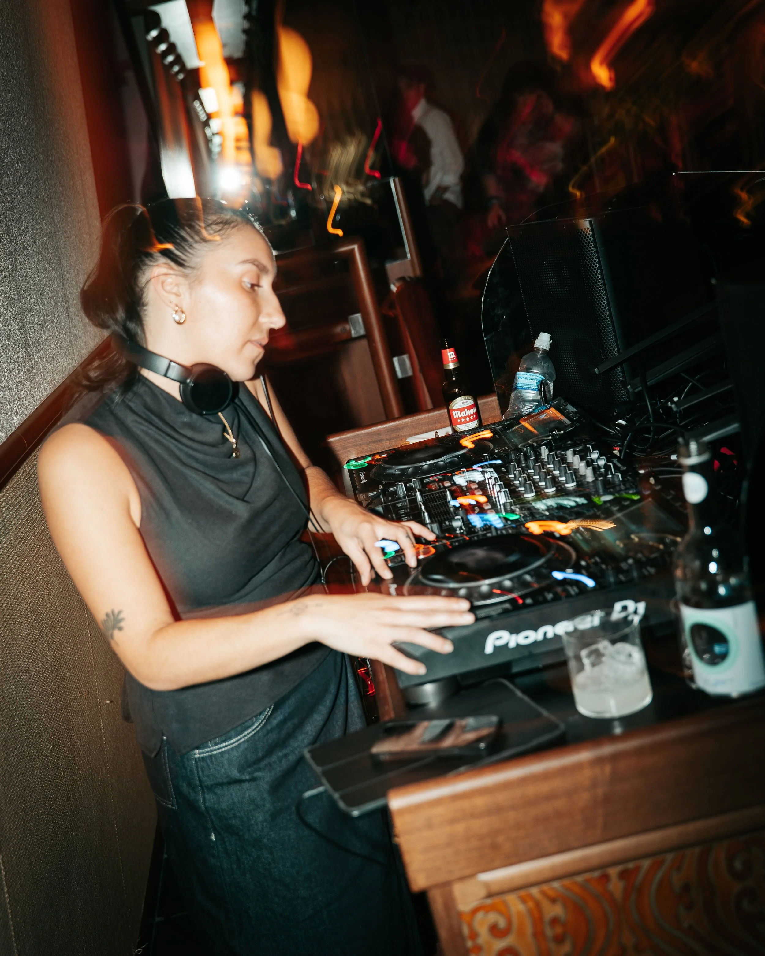 A female DJ with headphones around her neck, operating a Pioneer DJ controller in a dimly lit setting with blurred partygoers in the background.
