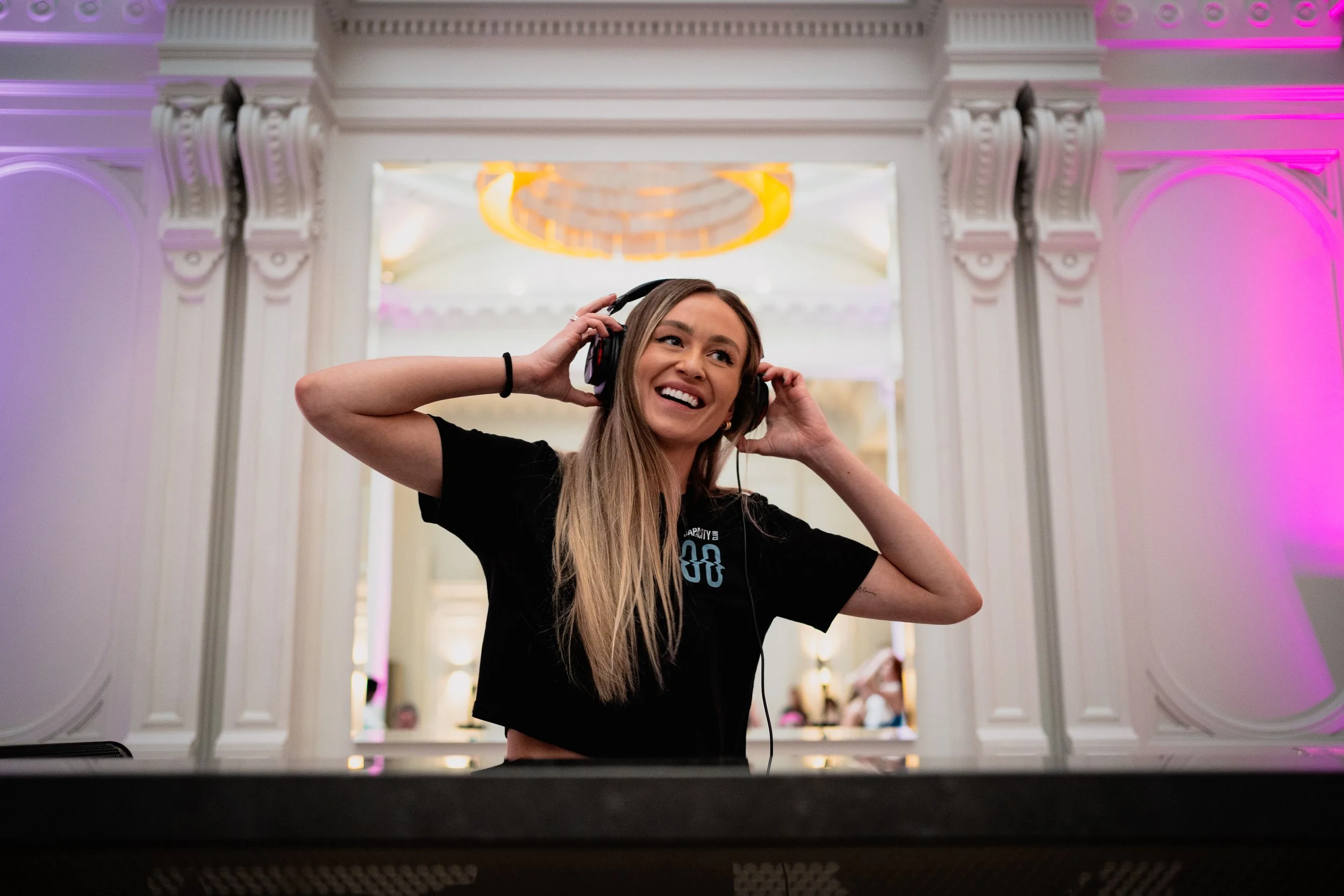 A young woman with long blonde hair, wearing a black T-shirt, smiling and adjusting her headphones while DJing at an event in an elegant ballroom with white ornate walls and pink and purple lighting.