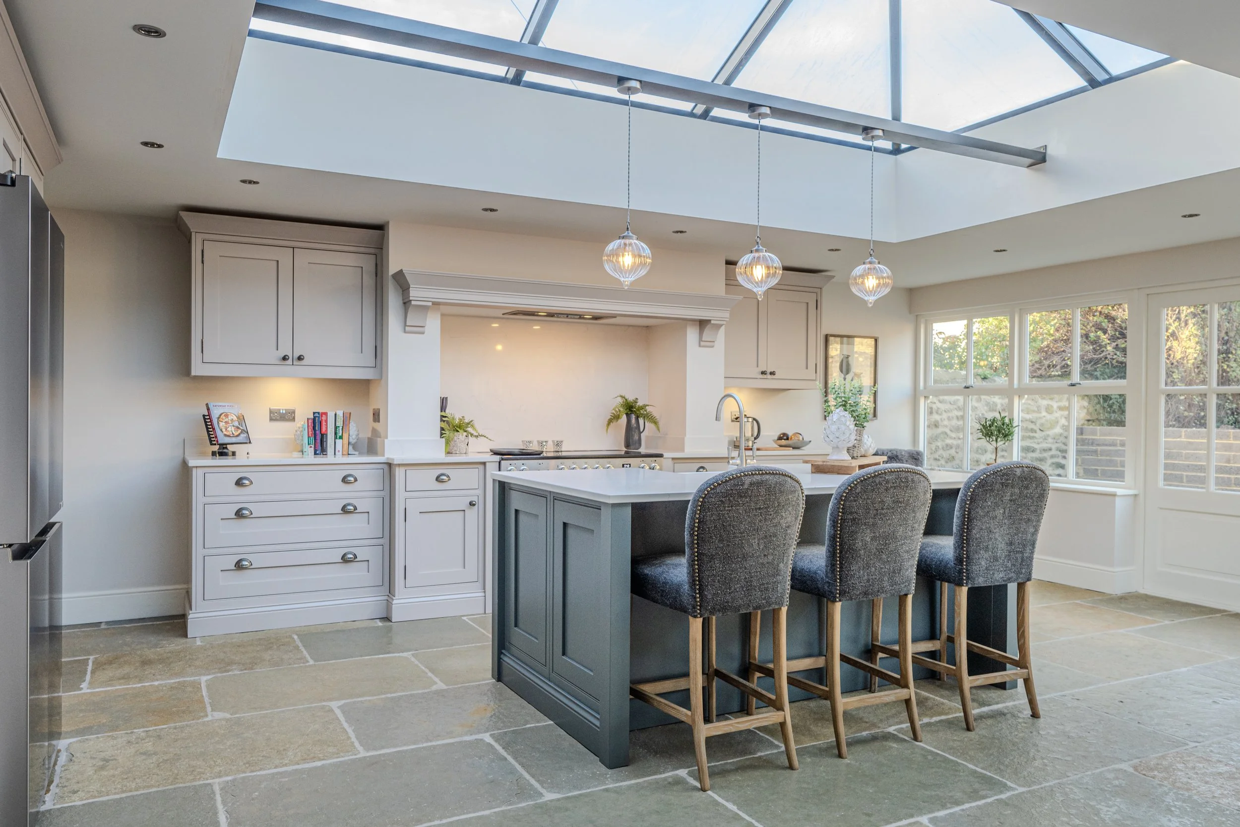 A bright modern kitchen with a large skylight overhead, featuring a central island with a sink and seating for three, surrounded by white cabinetry, large windows, and decorative plants.