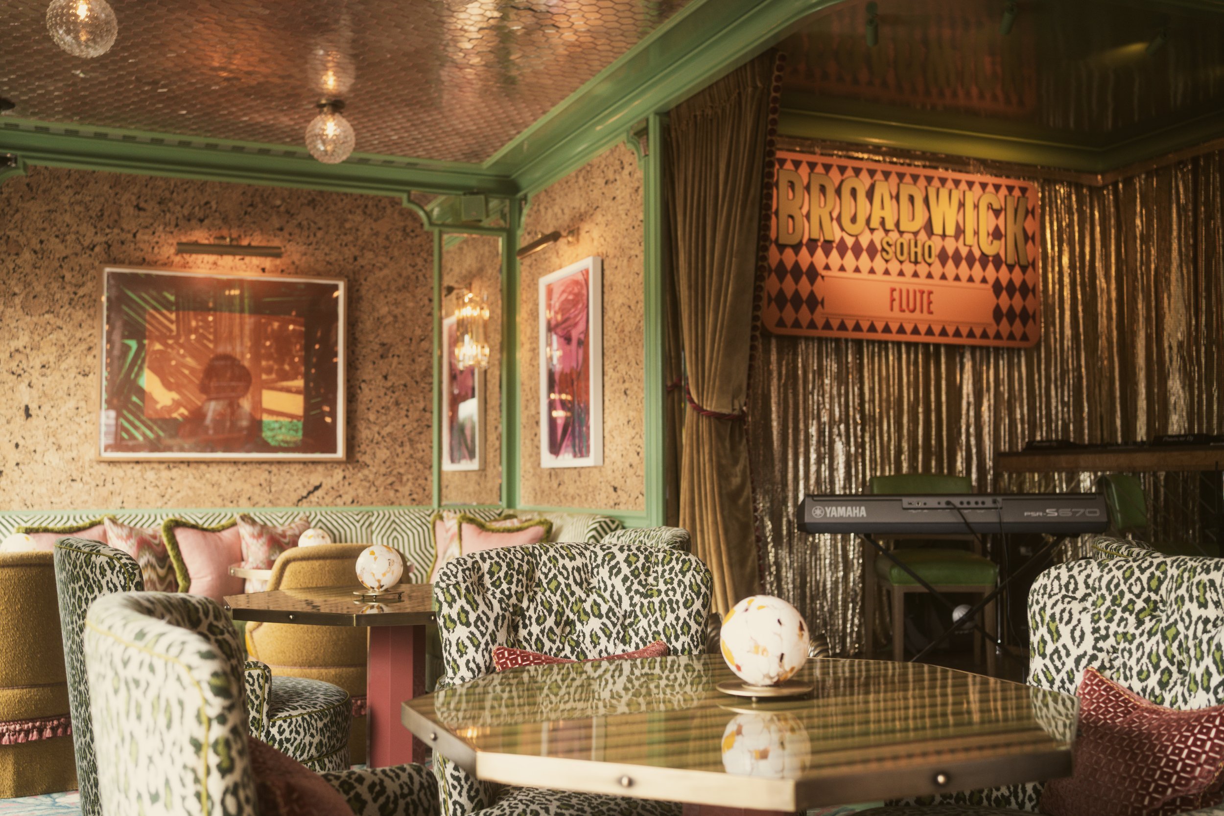 Interior of a stylish lounge area with leopard print chairs, pink pillows, gold decor, framed pictures on the wall, a sign reading 'Broadwick Soho Flute,' and musical keyboard on a stand.