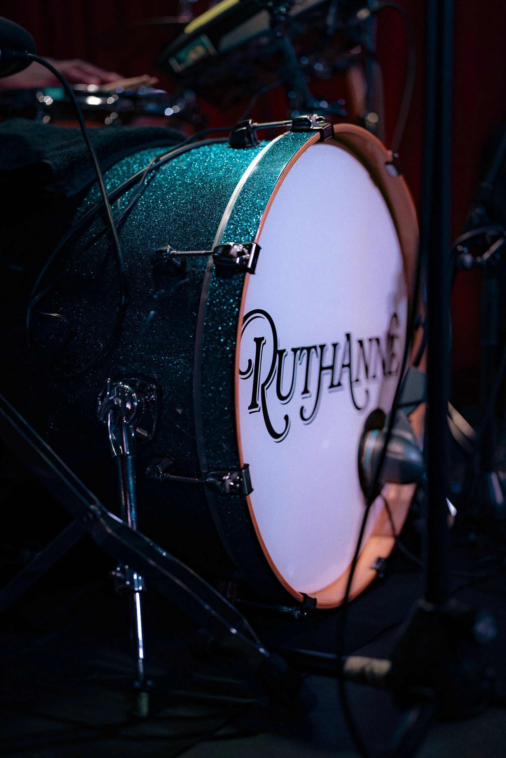 A close-up of a drum with a glittery teal finish and a white drumhead displaying the logo 'Ruthann' in black script, surrounded by musical equipment and cables.