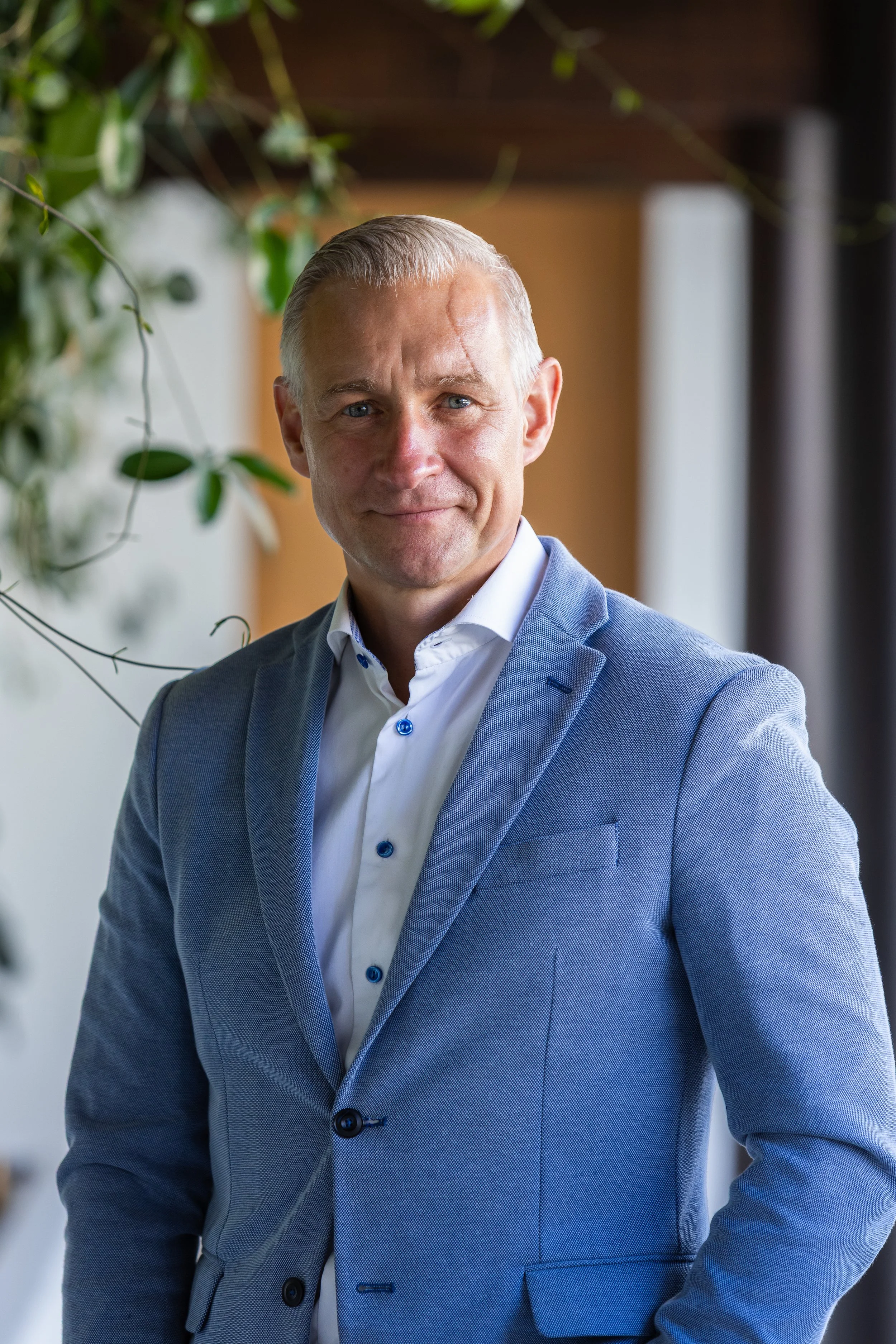 A middle-aged man with short gray hair and blue eyes, wearing a light blue blazer and white shirt, standing indoors with plants and a blurred background.