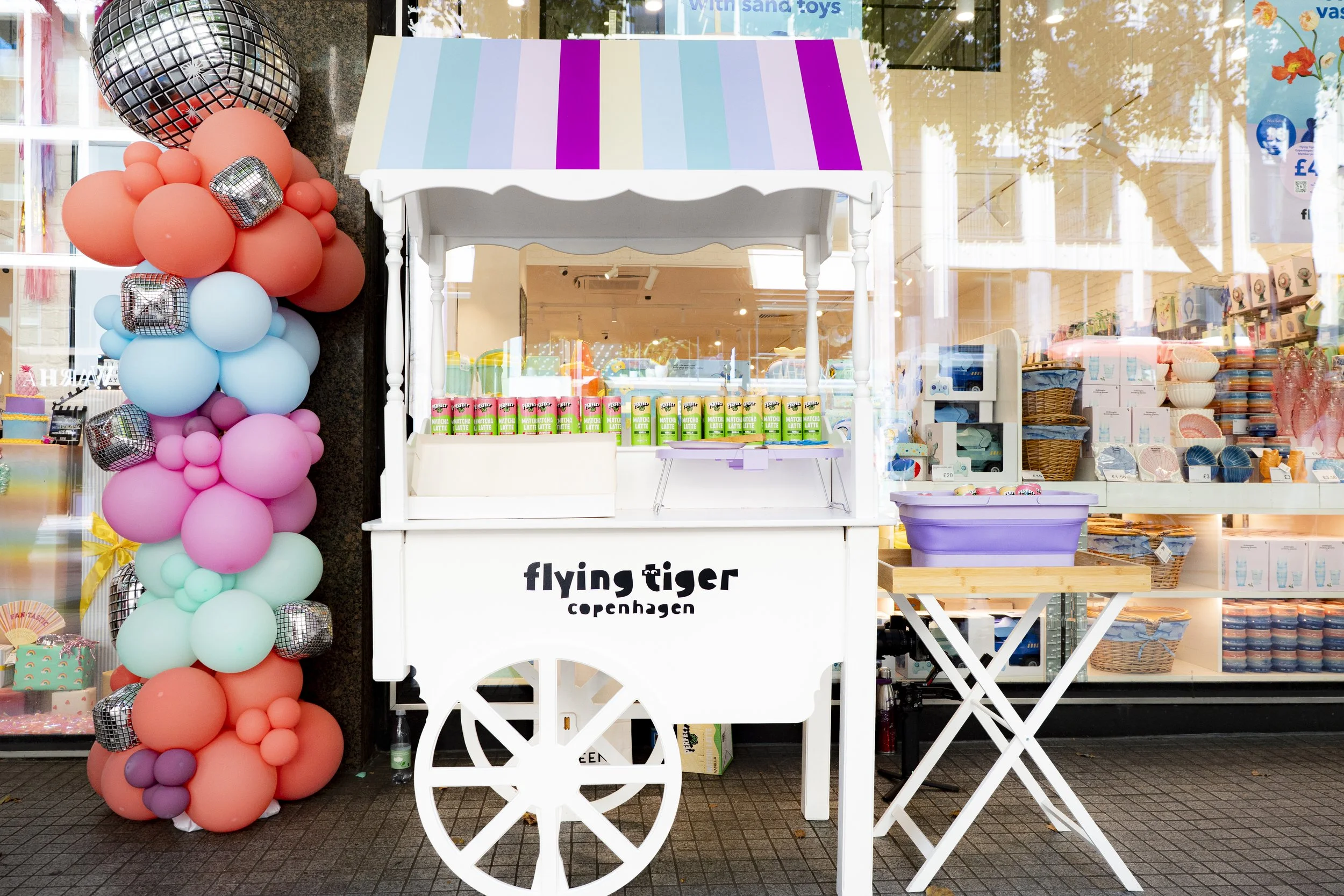 A white mobile cart with the words 'flying tiger Copenhagen' on it, set up outside a store. The cart has colorful cans on top and is decorated with pastel-colored balloons in shades of coral, turquoise, pink, and purple, with some small metallic ball