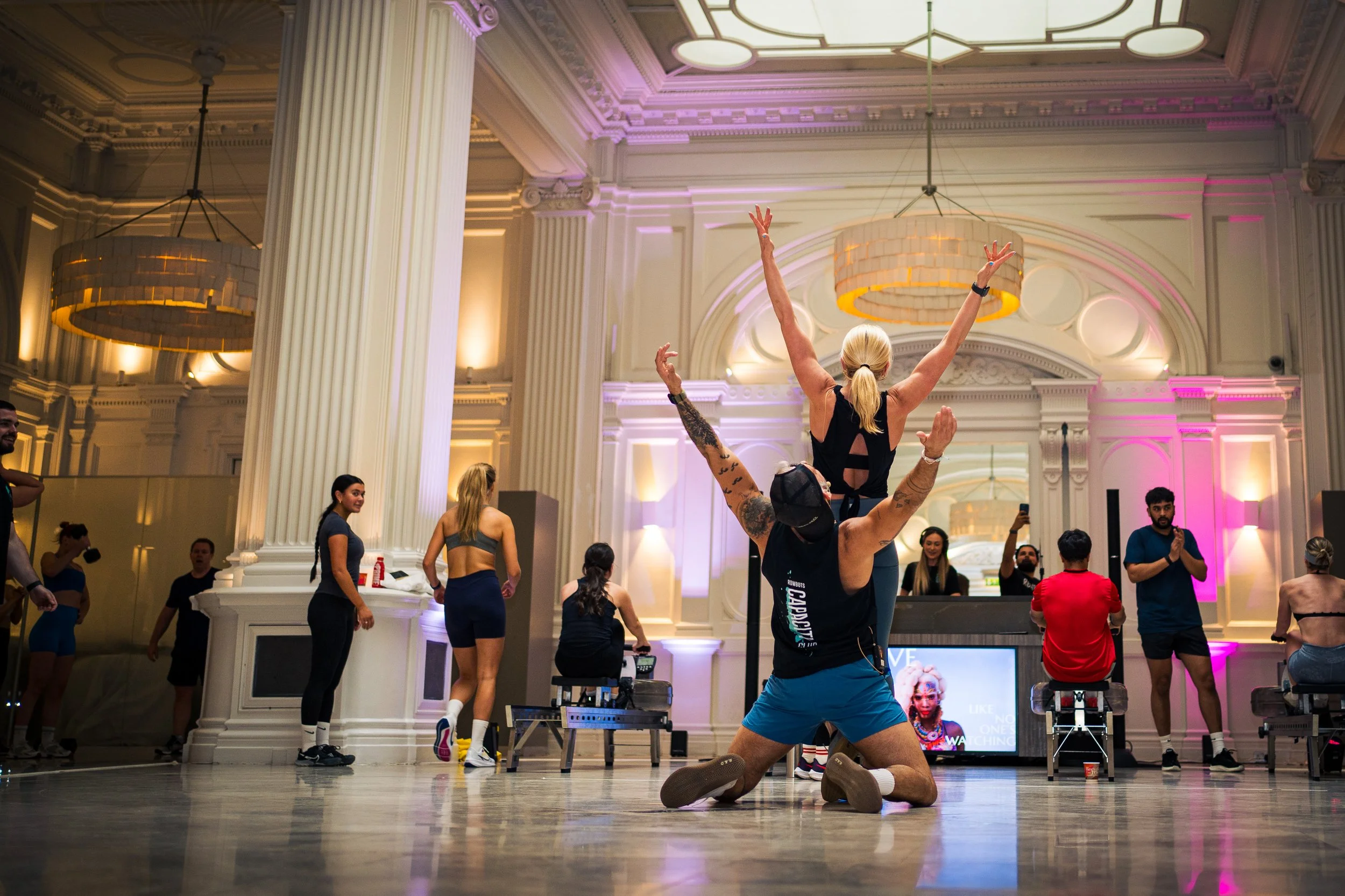 People dancing and taking photos in a grand hall with white pillars and purple lighting.