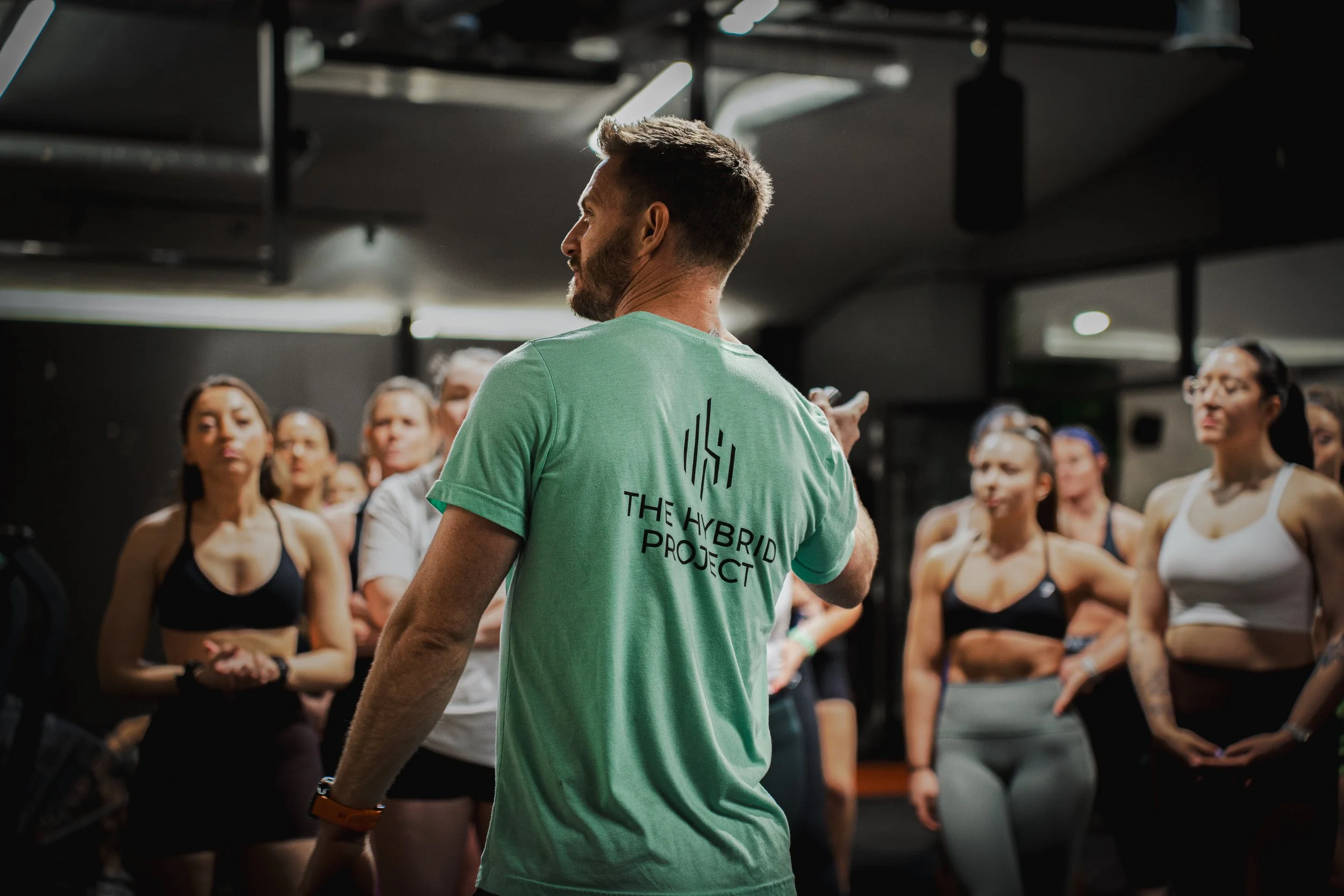 A male fitness instructor with a beard, wearing a green T-shirt labeled "The Hybrid Project," instructs a group of women exercising in a gym.