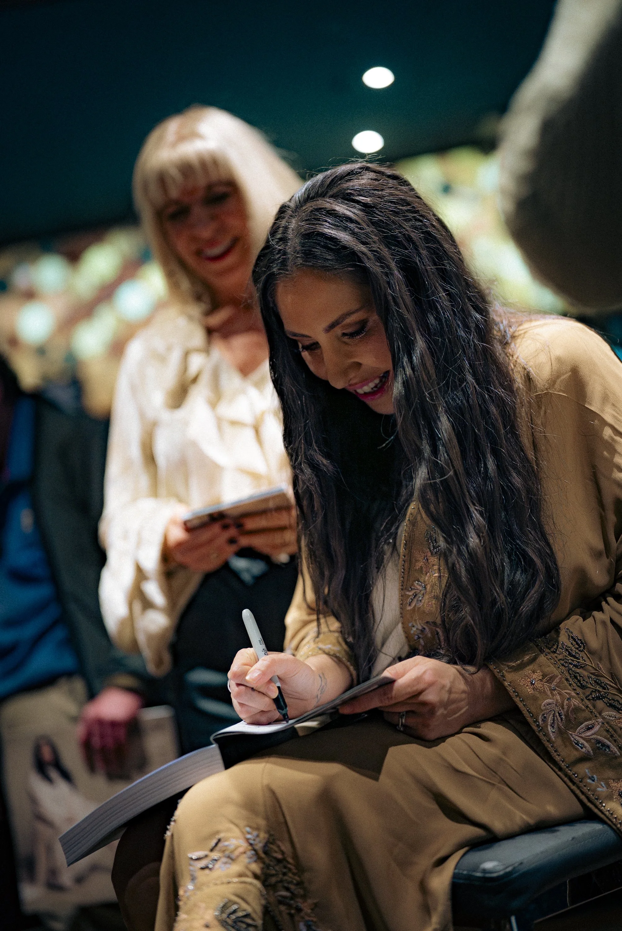 Two women, one with long dark hair and another with blonde hair, are smiling and signing autographs at an event.