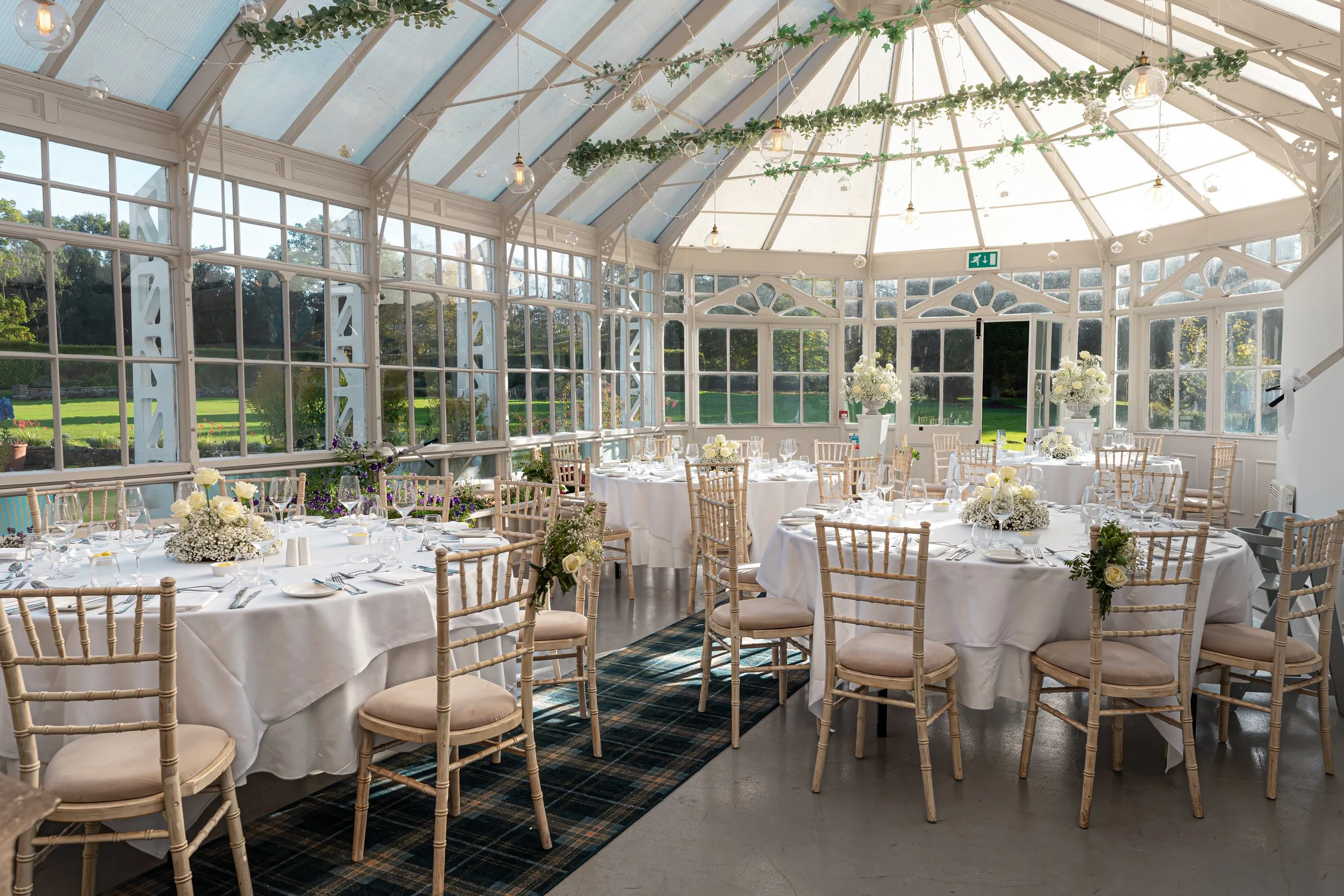 Elegant event space with round tables covered in white tablecloths, decorated with floral centerpieces, placed inside a glass-walled conservatory with greenery and natural light.