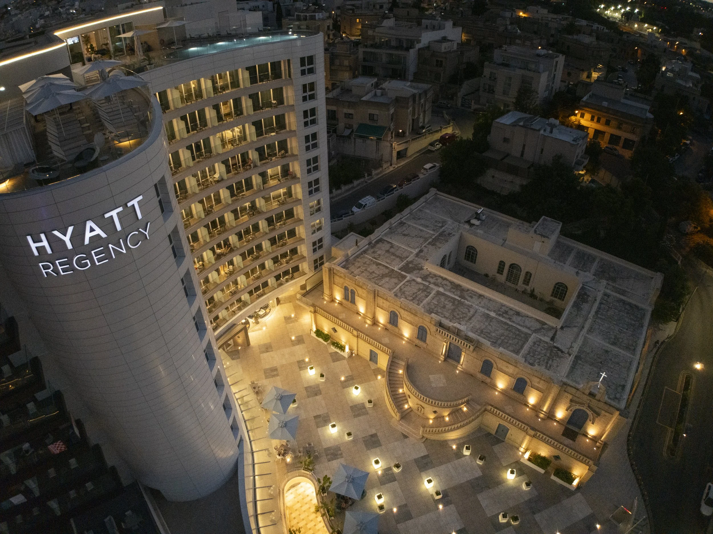 Aerial nighttime view of the Hyatt Regency hotel with a modern curved tower and multiple balconies, neighboring older buildings, and a well-lit courtyard with tables and umbrellas.