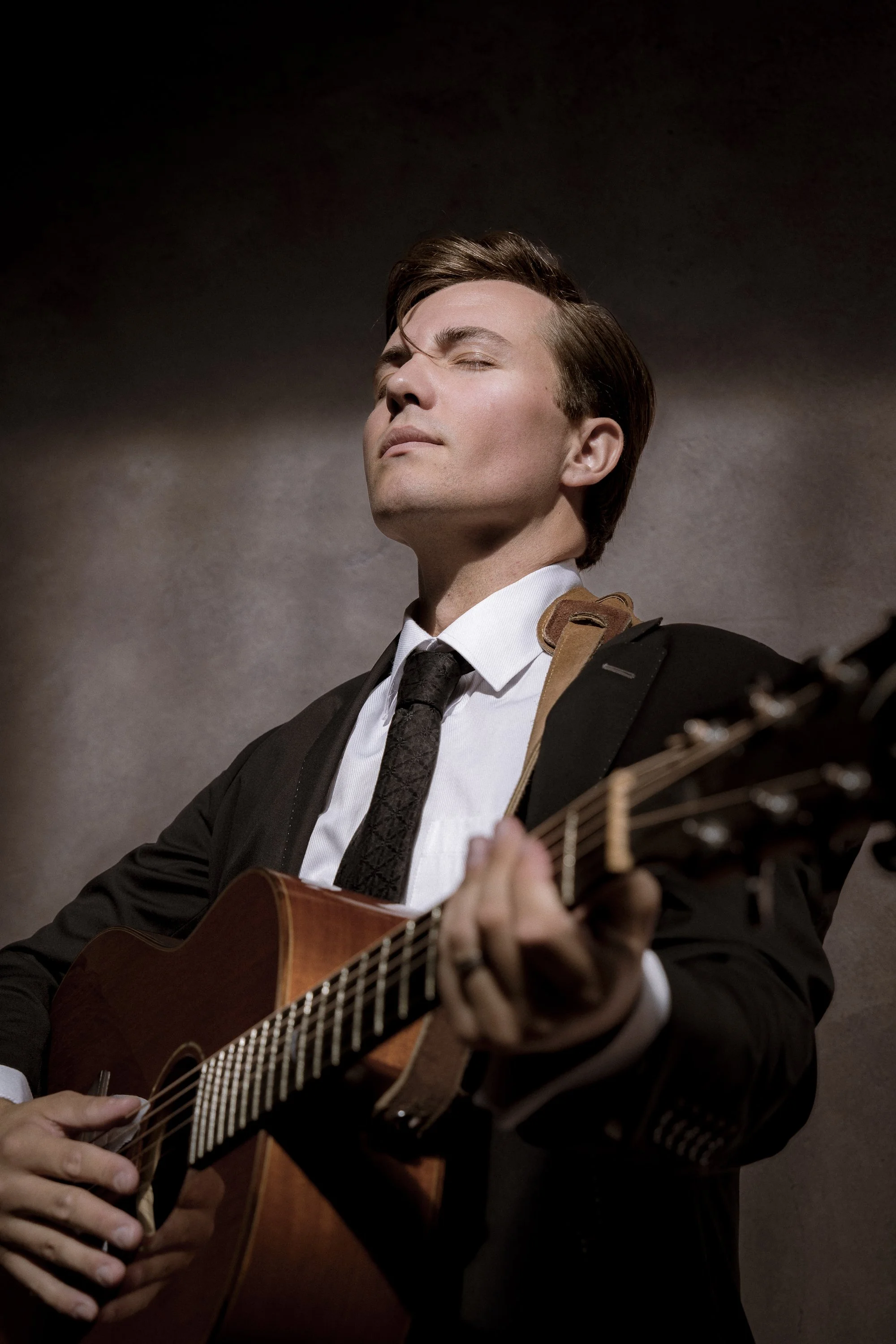A young man in a black suit playing an acoustic guitar with a focused and serene expression.