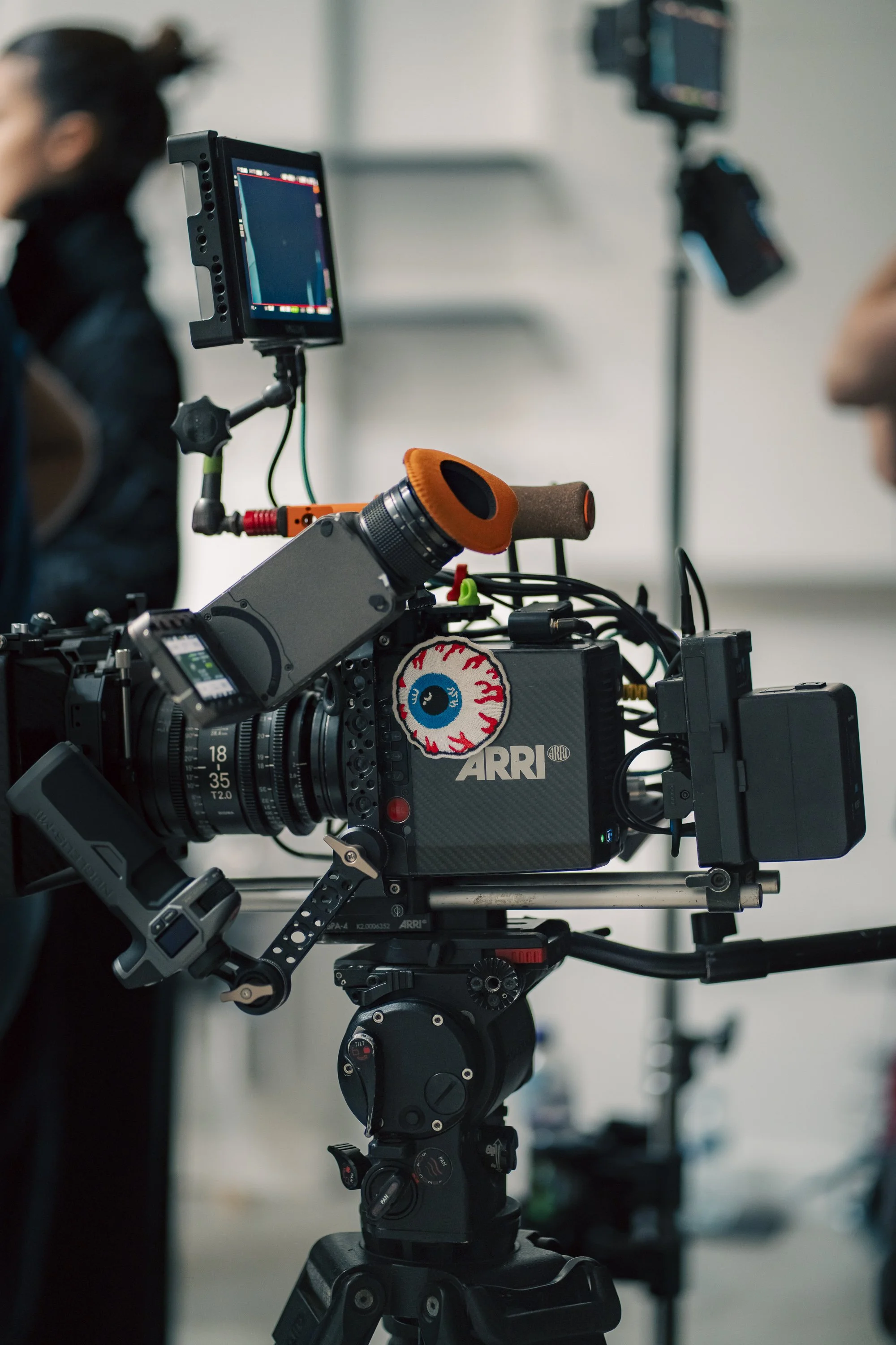 Professional ARRI cinema camera on a tripod with video monitor in a film or photography studio, blurred background of crew members and equipment.