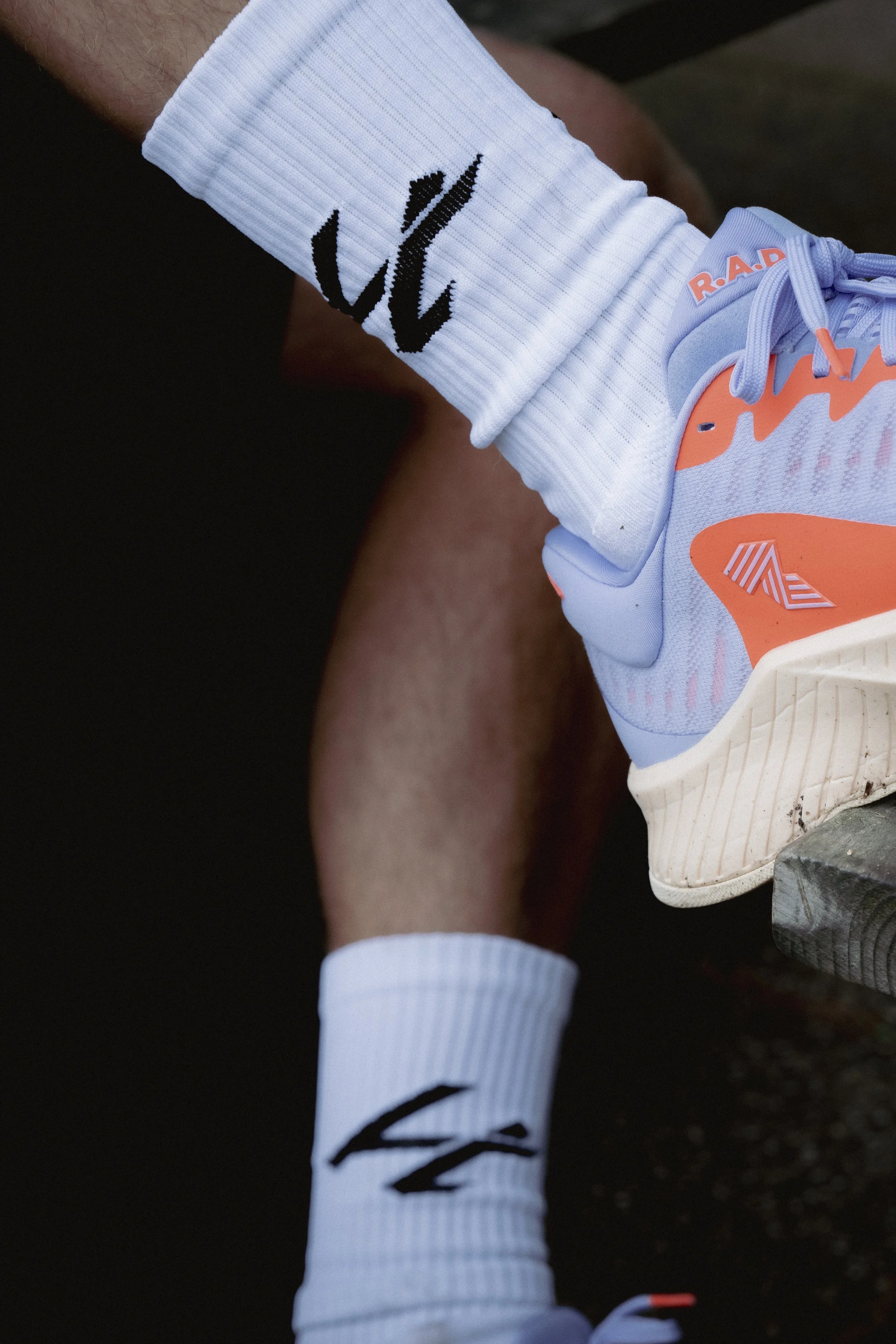 Close-up of a person's legs, one bent and one extended, wearing white Nike socks with black logos and pastel-colored running shoes with orange and purple accents, on a dark surface.