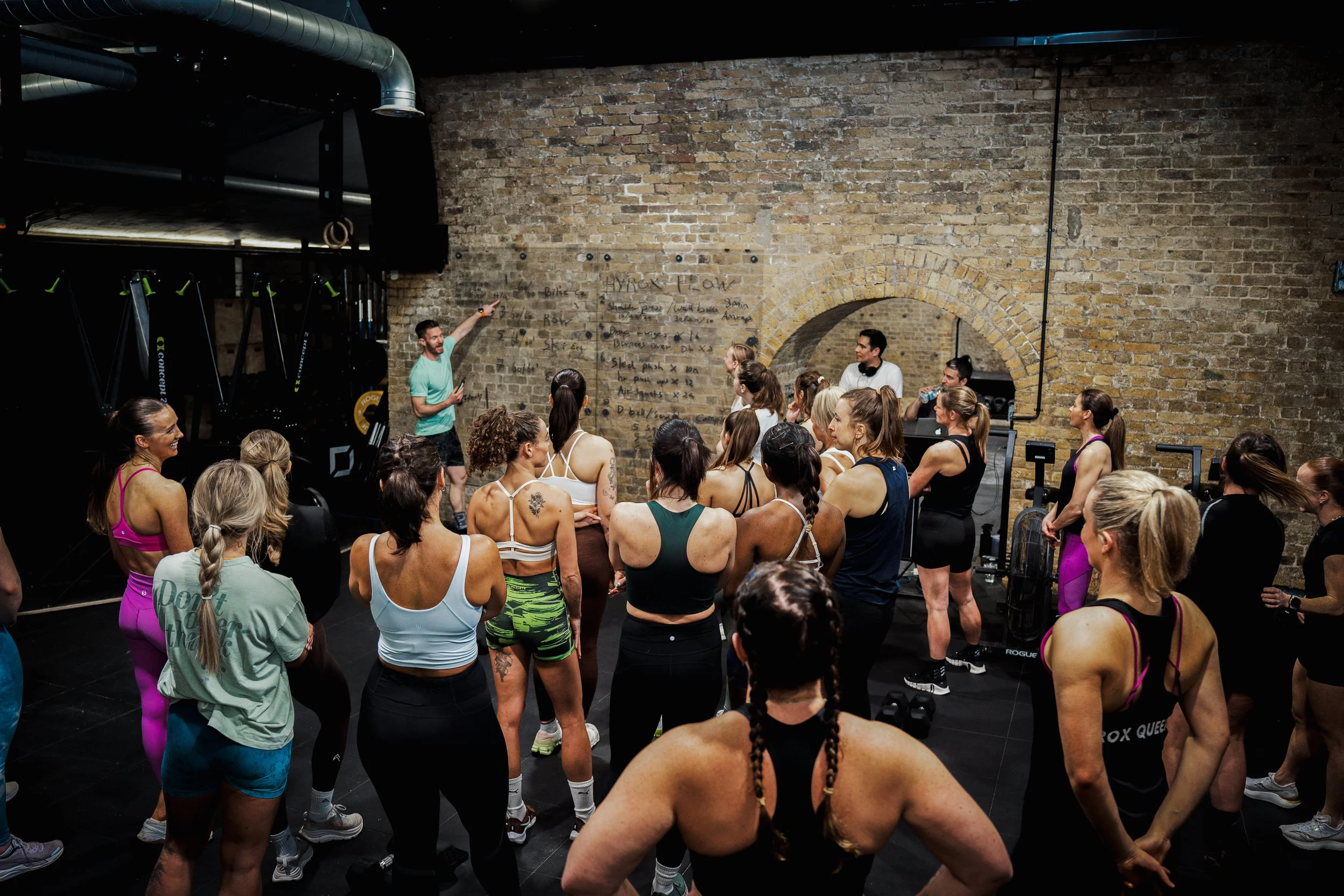 Group of women attending a fitness class in a brick-walled gym with a male instructor at the front