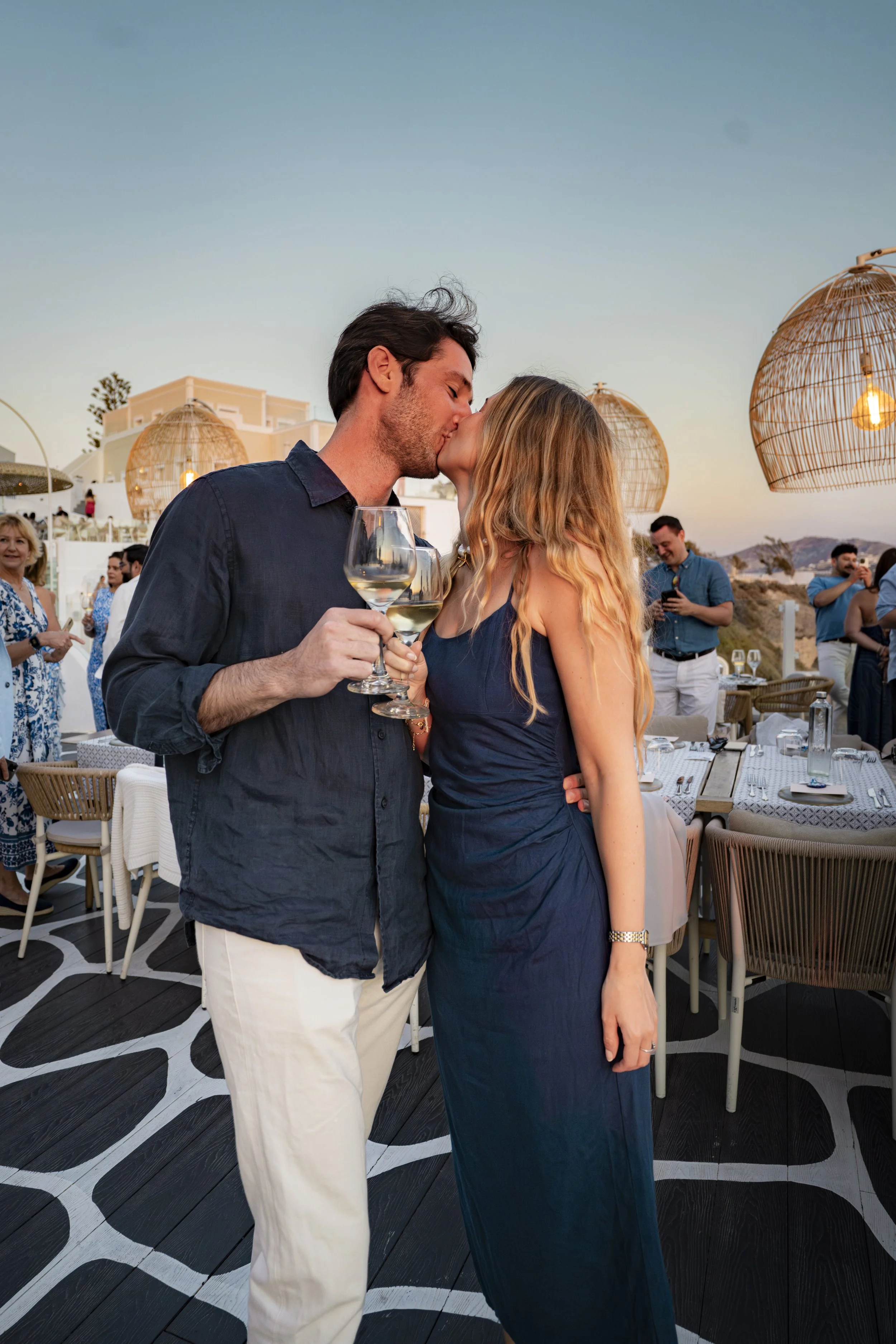 A couple shares a kiss at a romantic outdoor dinner event with string lights, while holding wine glasses, during sunset.