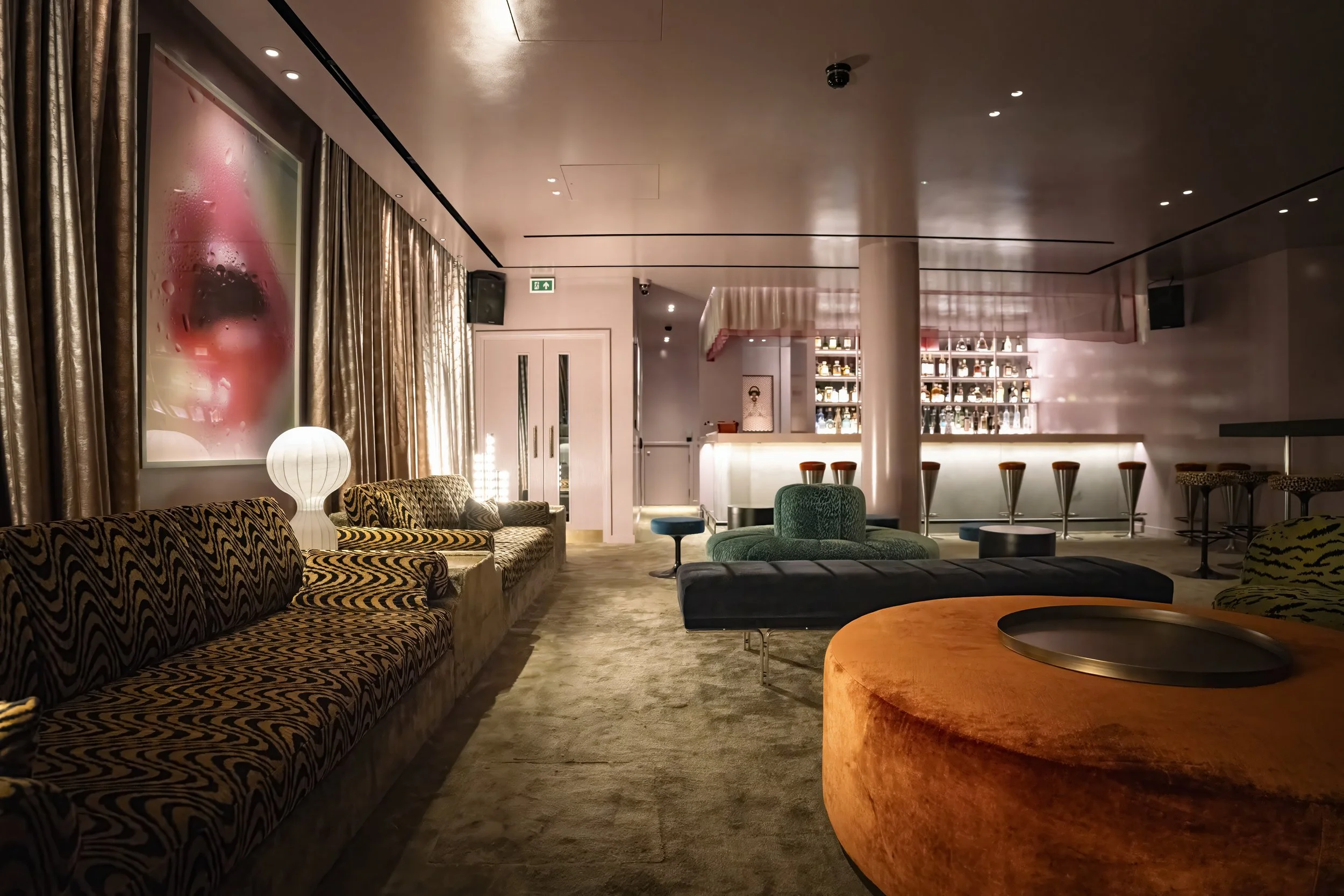 Interior of a stylish lounge bar with patterned couches, a bar counter with stools, and soft ambient lighting.