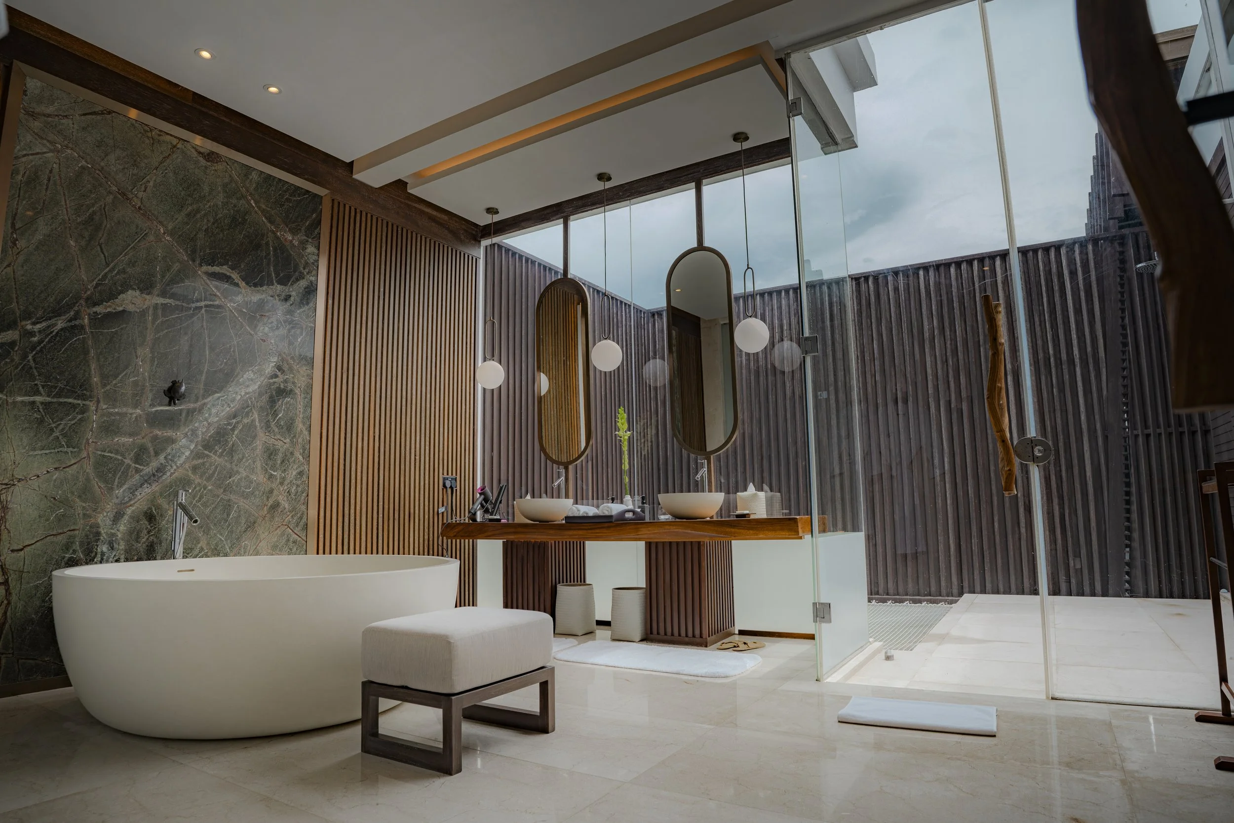 Modern bathroom with free-standing bathtub, double vanity with oval mirrors, and an outdoor shower area enclosed by wooden fencing.