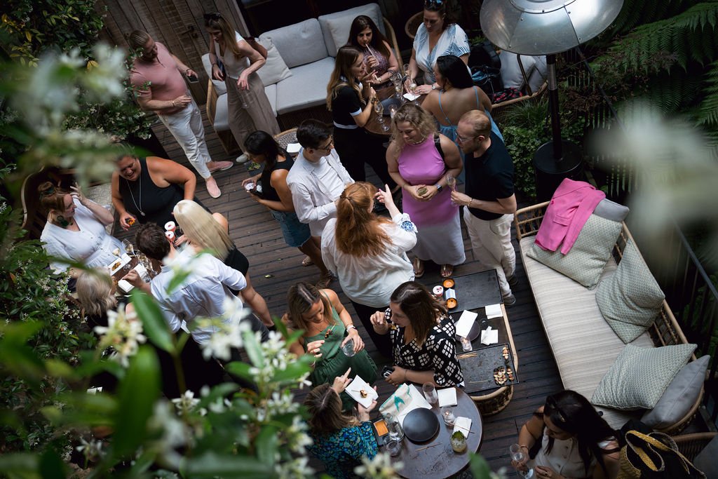Group of people socializing at a backyard party with lush greenery, outdoor furniture, and drinks.