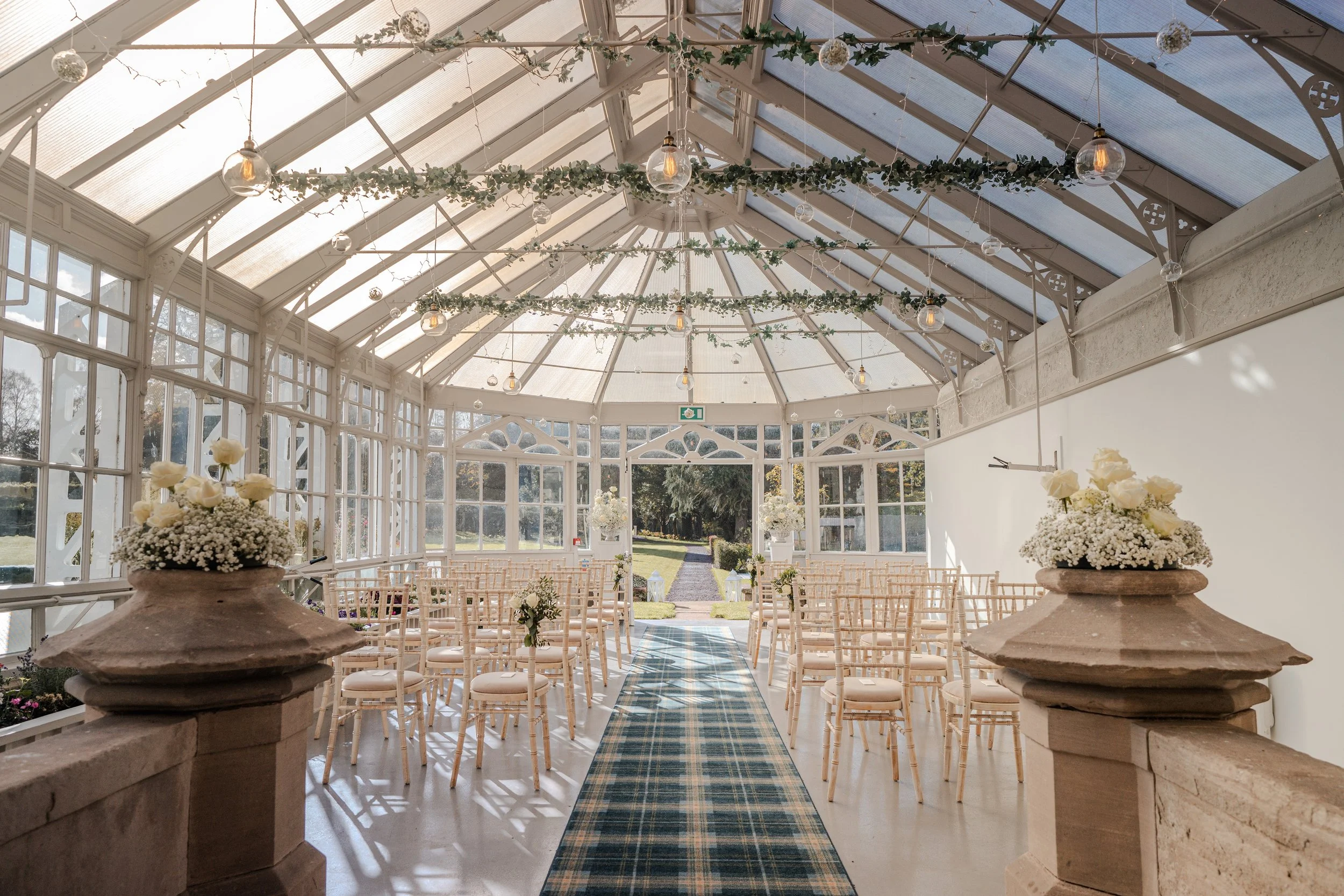 A bright, elegant indoor wedding ceremony setup inside a glass conservatory with white walls and ceiling, decorated with greenery and hanging light bulbs, with rows of beige chairs, floral arrangements, and a blue checkered aisle runner leading to an