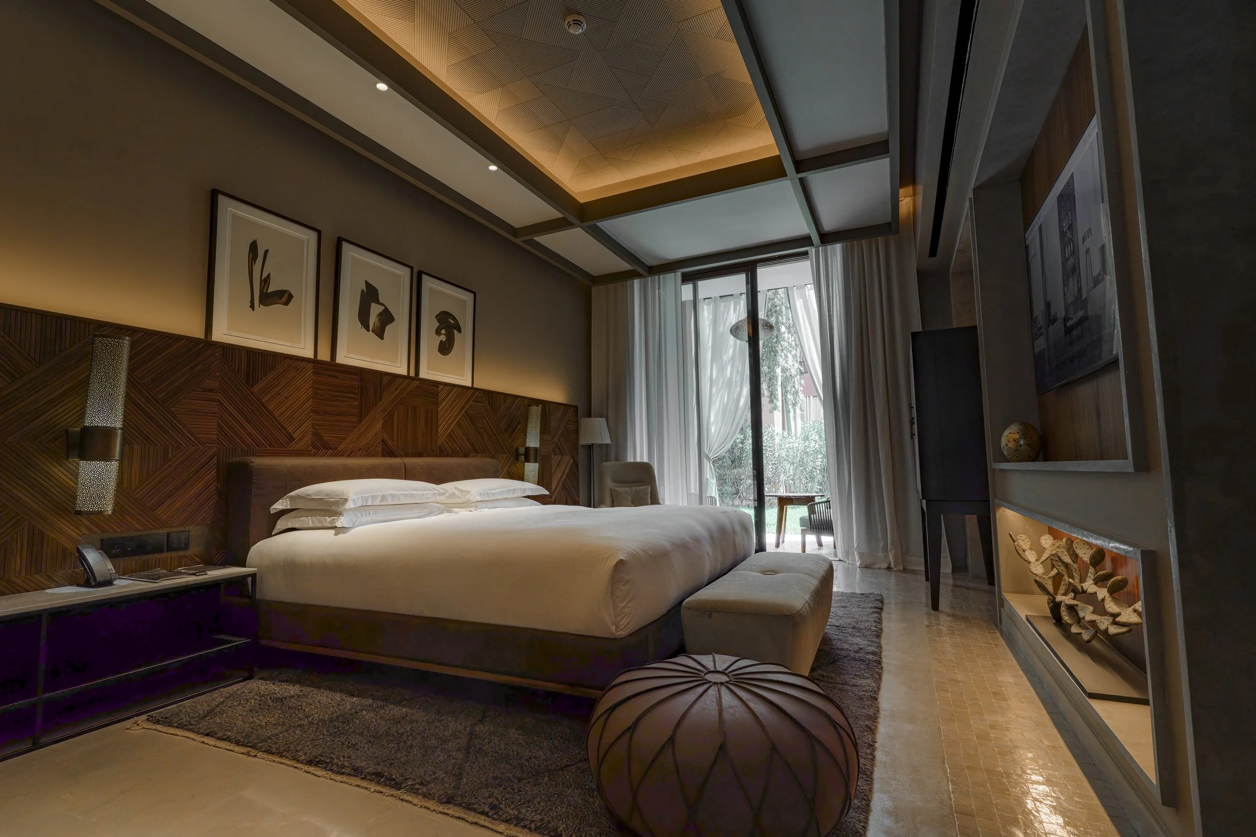 A modern elegant hotel bedroom with a large bed, bedside table, artwork above the bed, a sitting chair, and a view of a balcony with outdoor furniture.