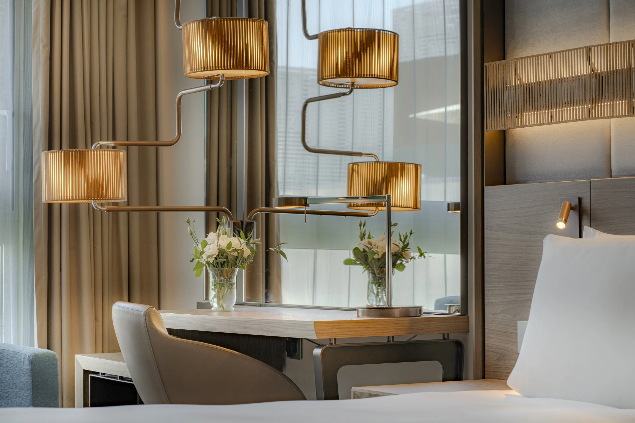 Modern hotel room with a bed, side table, and decorative lighting fixtures on a wooden headboard and a table with vases of white flowers.