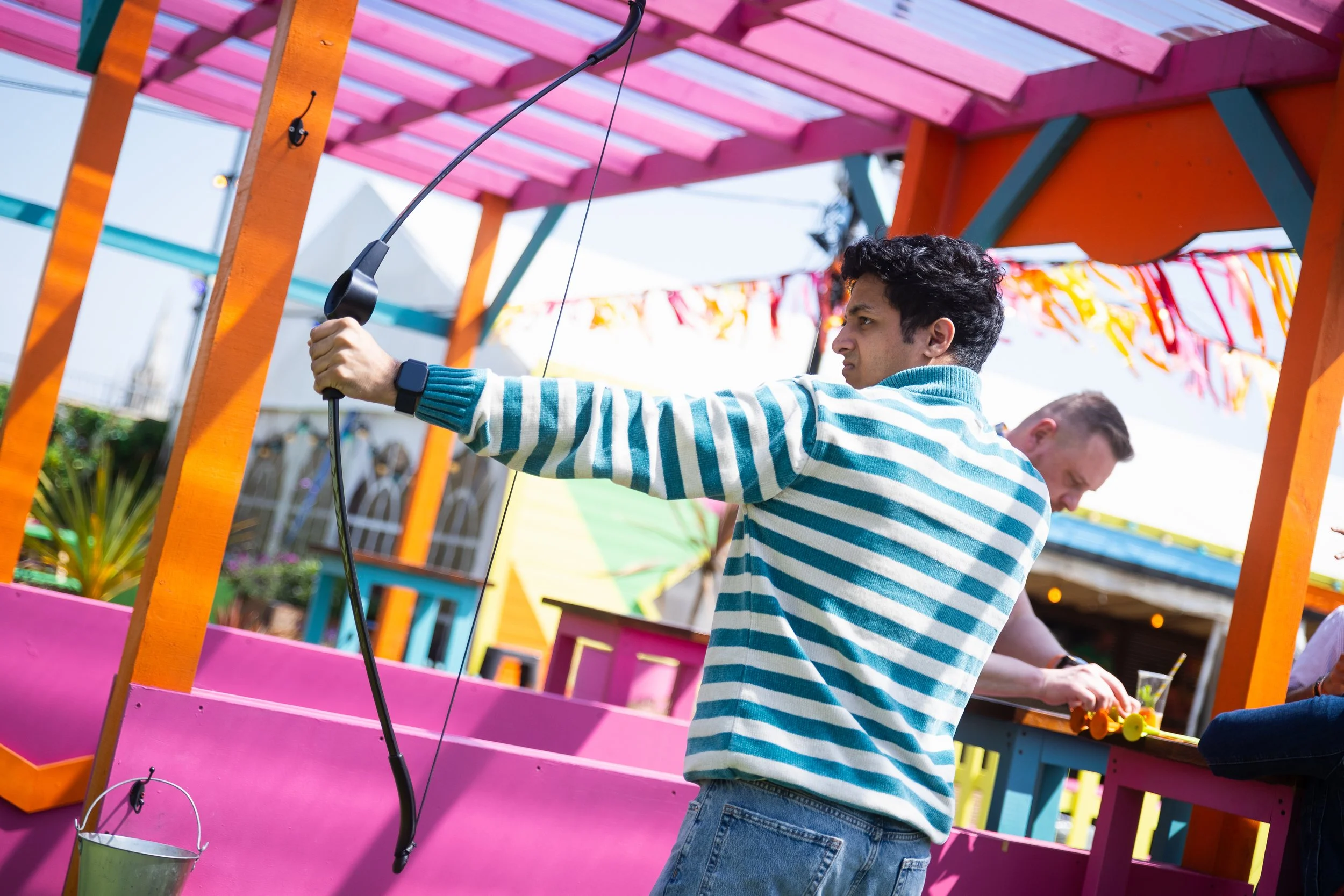 A man playing a game similar to archery at a colorful outdoor carnival or fair, with bright pink and orange decorations and others in the background.
