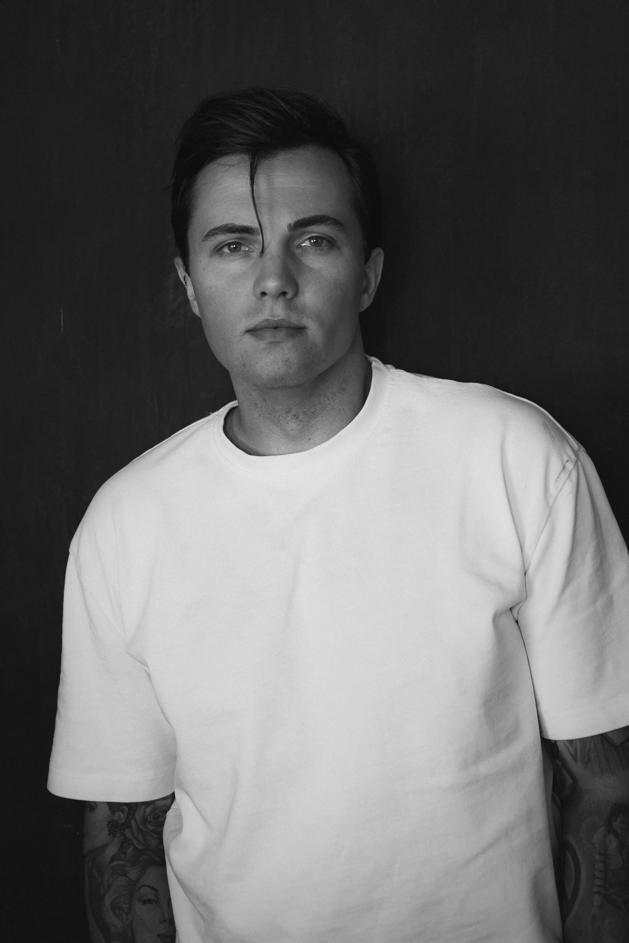 Black and white portrait of a young man with dark hair, wearing a plain white t-shirt, with tattoos visible on his arms, standing against a dark background.