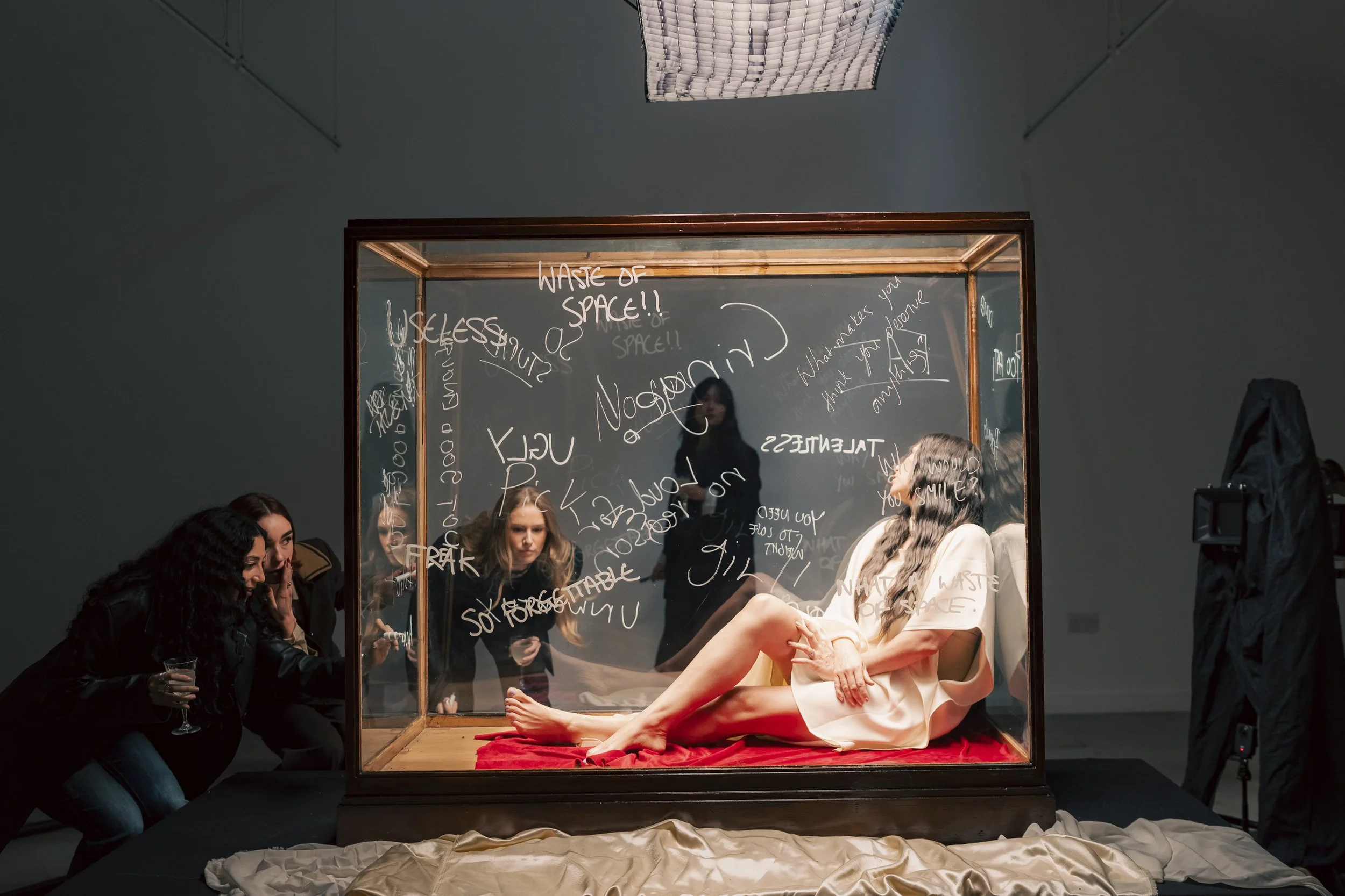 A woman in a white dress lying inside a glass display case with red fabric at the bottom, surrounded by people observing and writing on the glass, in an art gallery or exhibition setting.