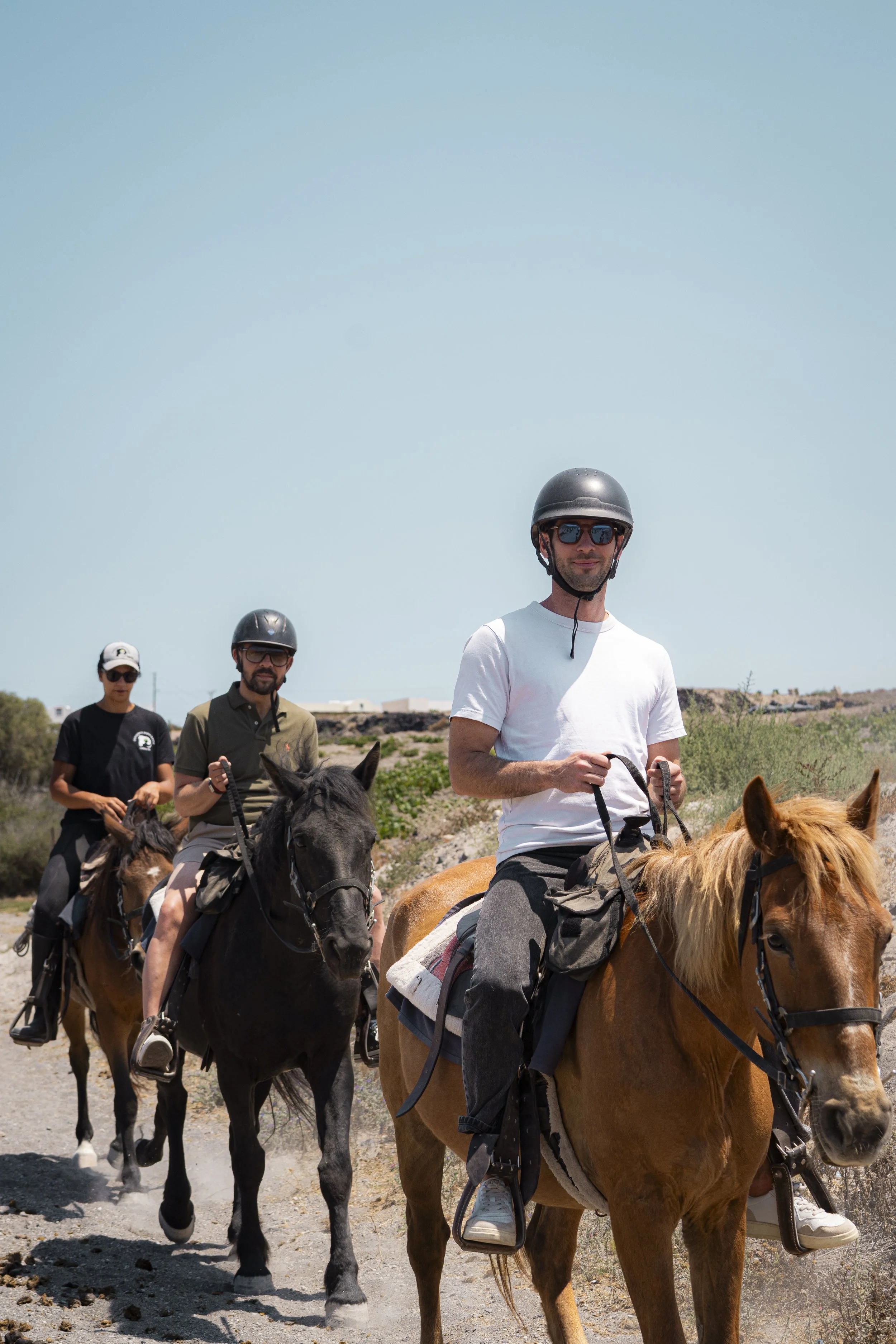 Three people riding horses on a trail under a clear blue sky, wearing helmets and casual clothing.