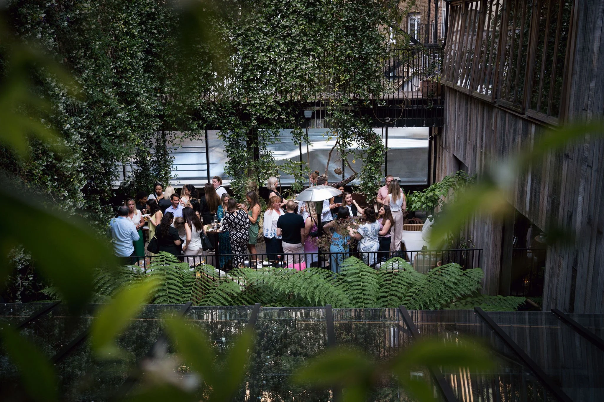 Group of people gathered outdoors on a patio surrounded by greenery and trees, with some holding drinks and engaging in conversation, in a garden-like setting.