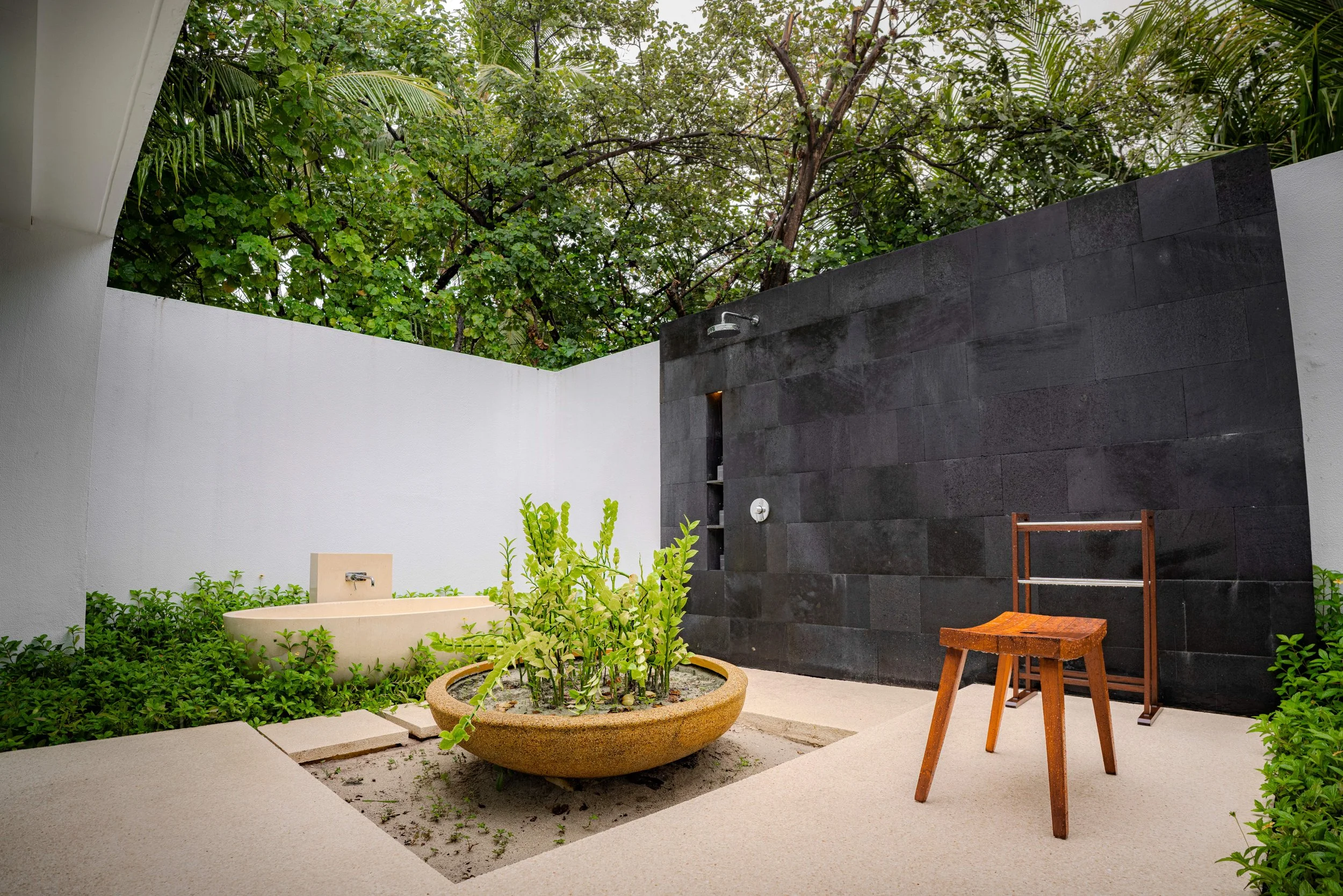 Modern outdoor shower area with black tiled wall, built-in shower, a wooden stool, and a large planter with green plants. The area is enclosed by white walls and surrounded by lush trees.