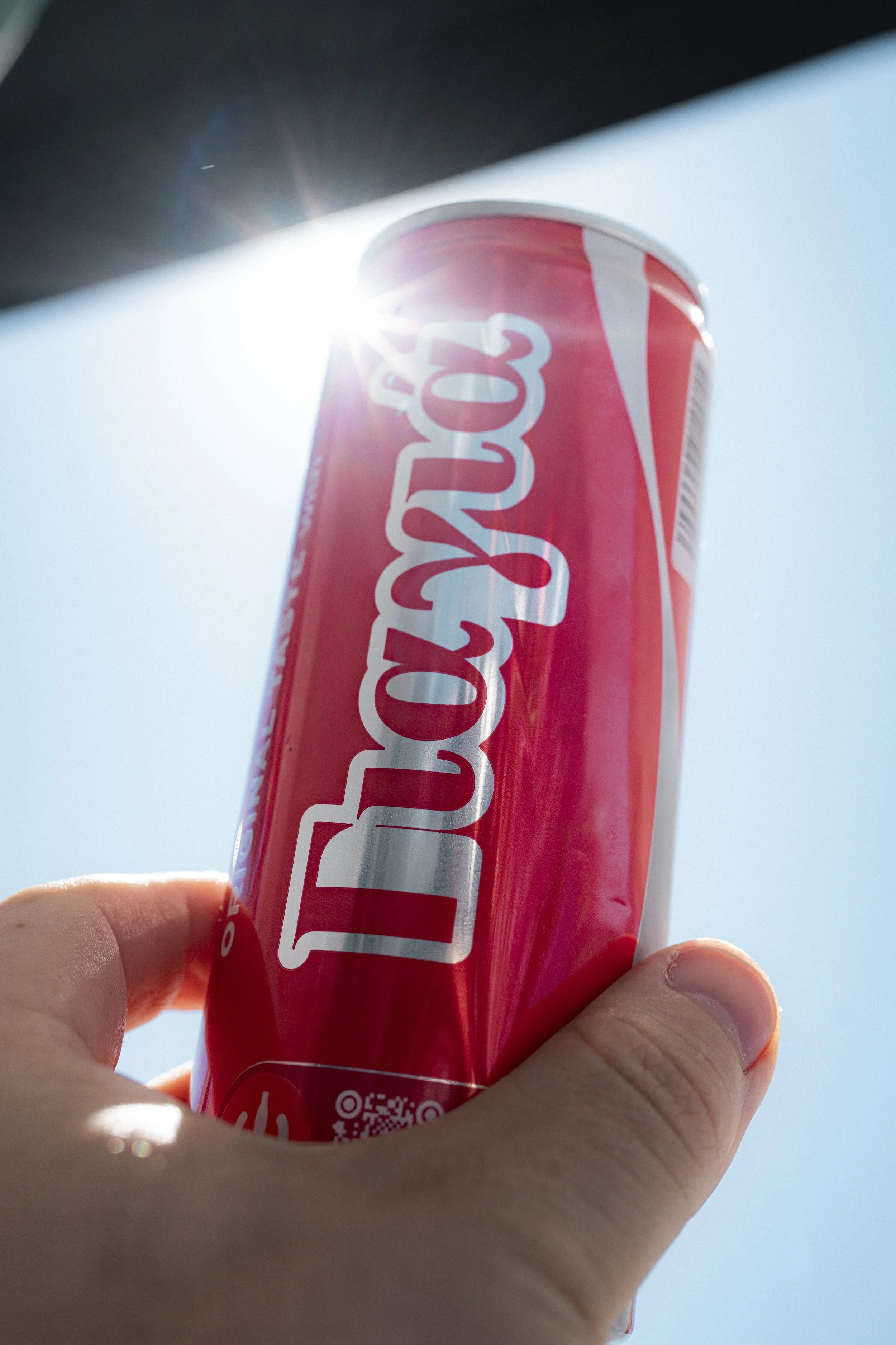 A hand holding a red and white twist-up can of Coca-Cola against a bright sky with sunlight shining behind it.