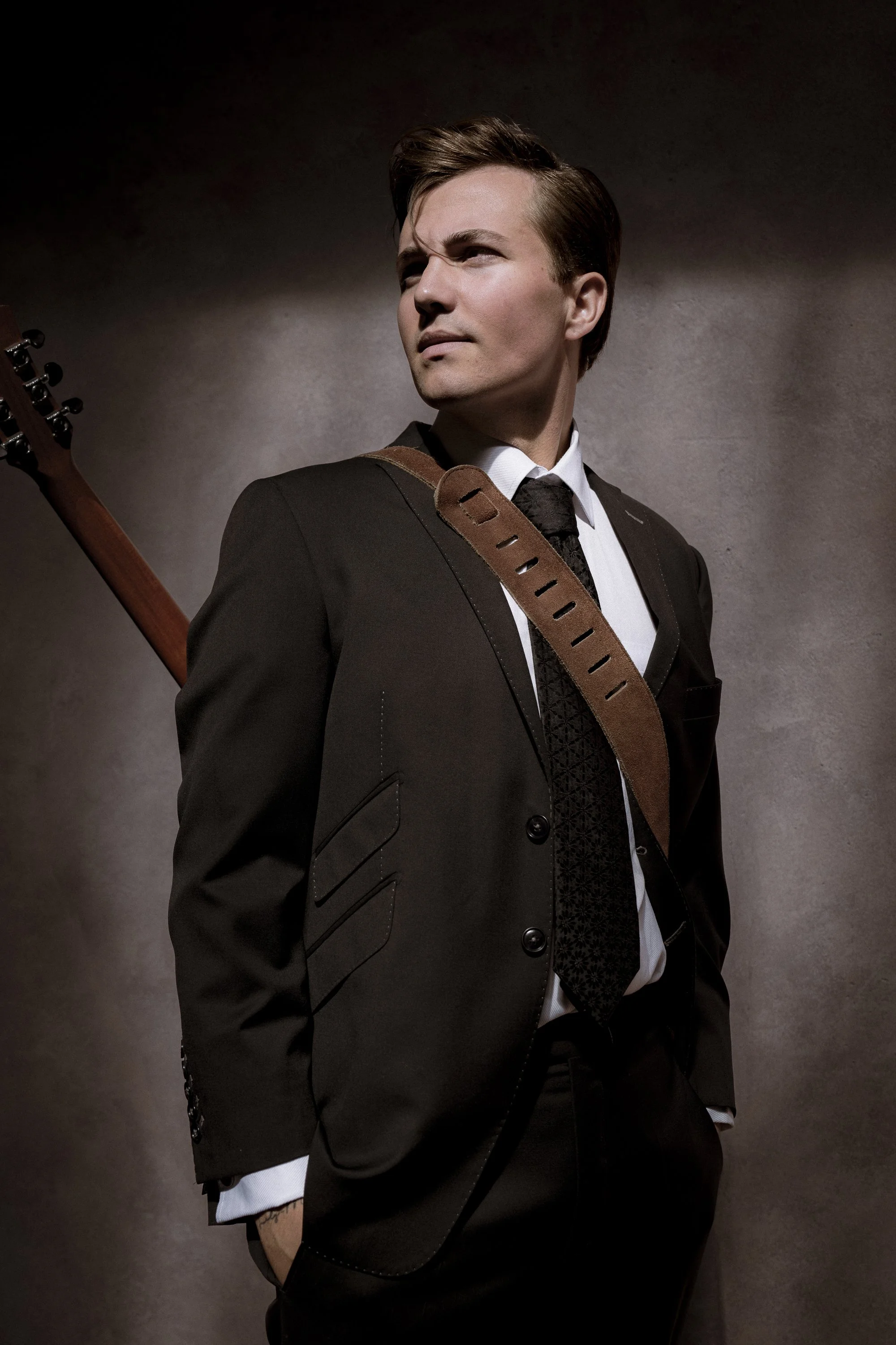 A man in a black suit with a white shirt and patterned black tie, standing with his hands in his pockets, wearing a guitar strap over his shoulder, and looking to the side against a plain background.