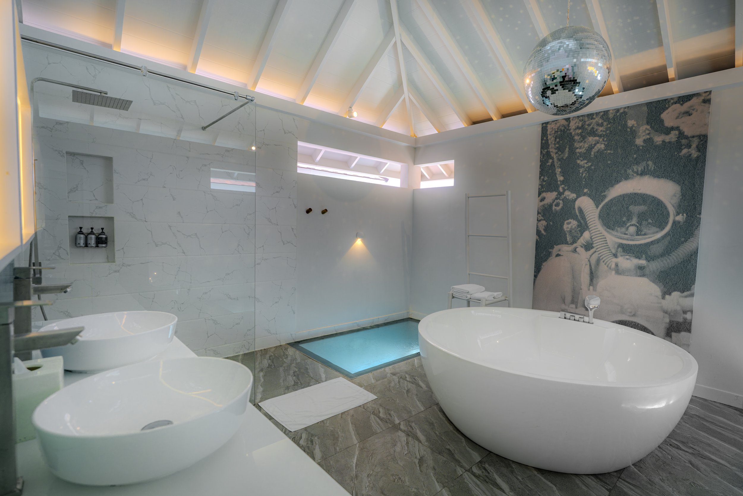 Modern bathroom with a white freestanding bathtub, a glass-enclosed shower with marble tiles, twin vessel sinks, a metallic disco ball hanging from the vaulted ceiling, and a large underwater-themed mural on the wall.