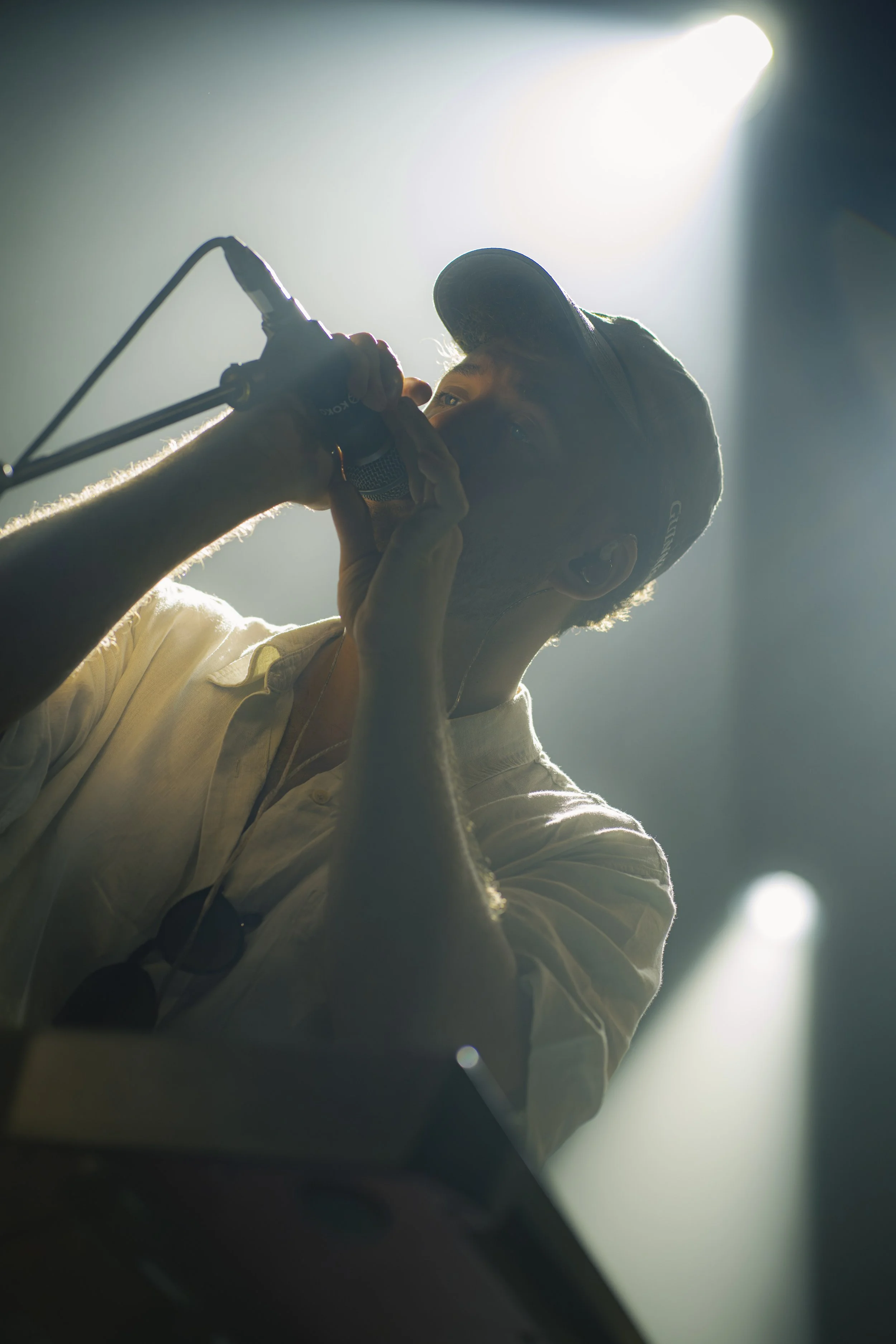 A man wearing a cap and white shirt singing into a microphone on stage with bright lighting.