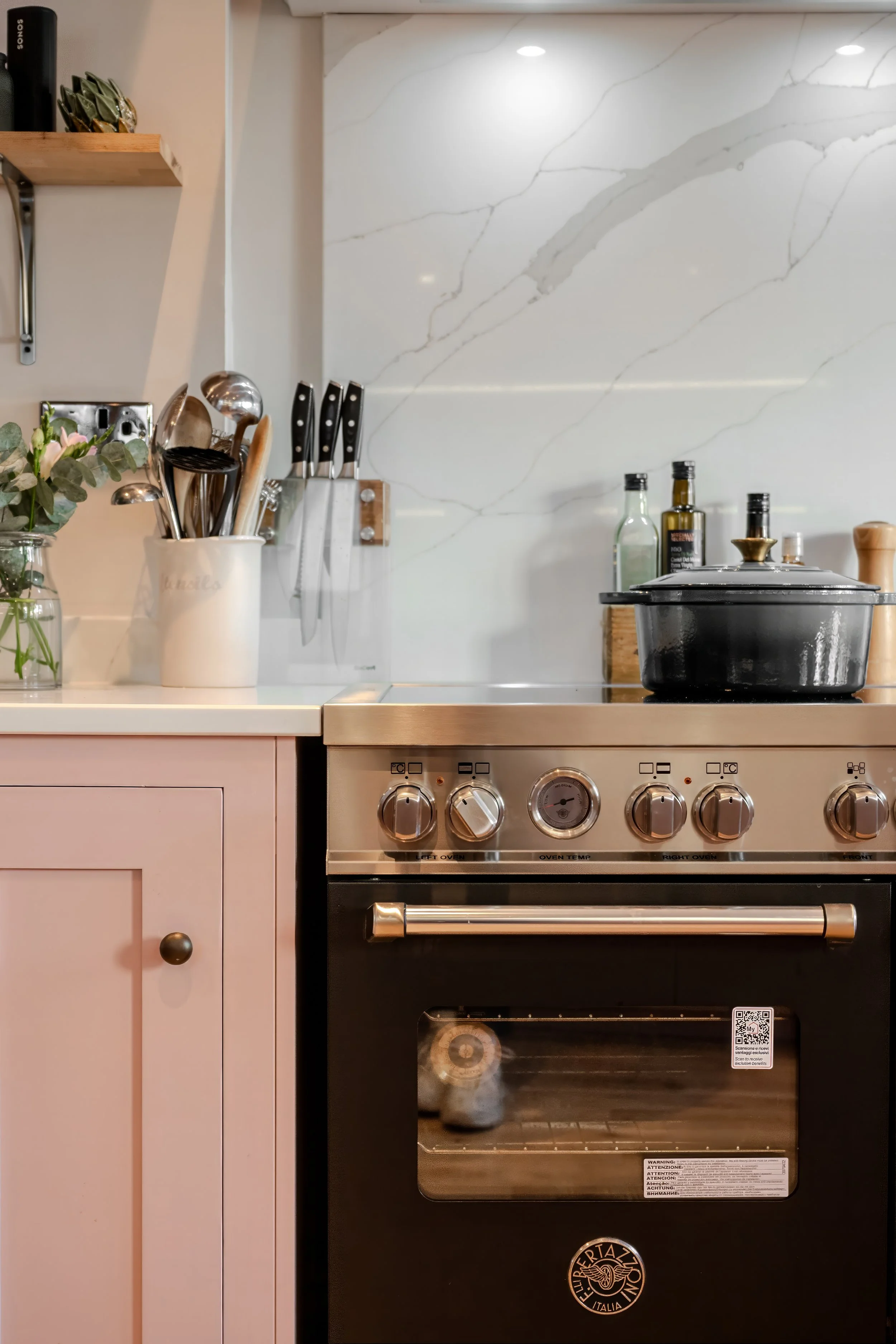 A modern kitchen with a black oven, white countertop, pink cabinetry, and a marble backsplash. Kitchen utensils are in a white holder, and there are bottles and a black pot on the stove.