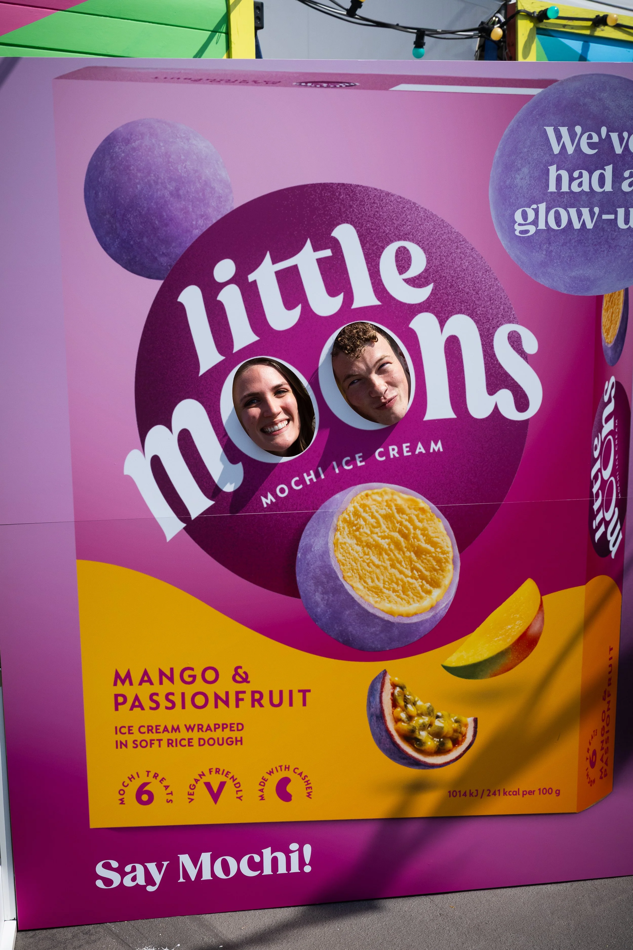Signboard advertising 'Little Moons' mochi ice cream with mango and passionfruit flavor, showing two people with faces in cutouts in the sign, surrounded by images of mango and passionfruit, with colorful decorations in the background.