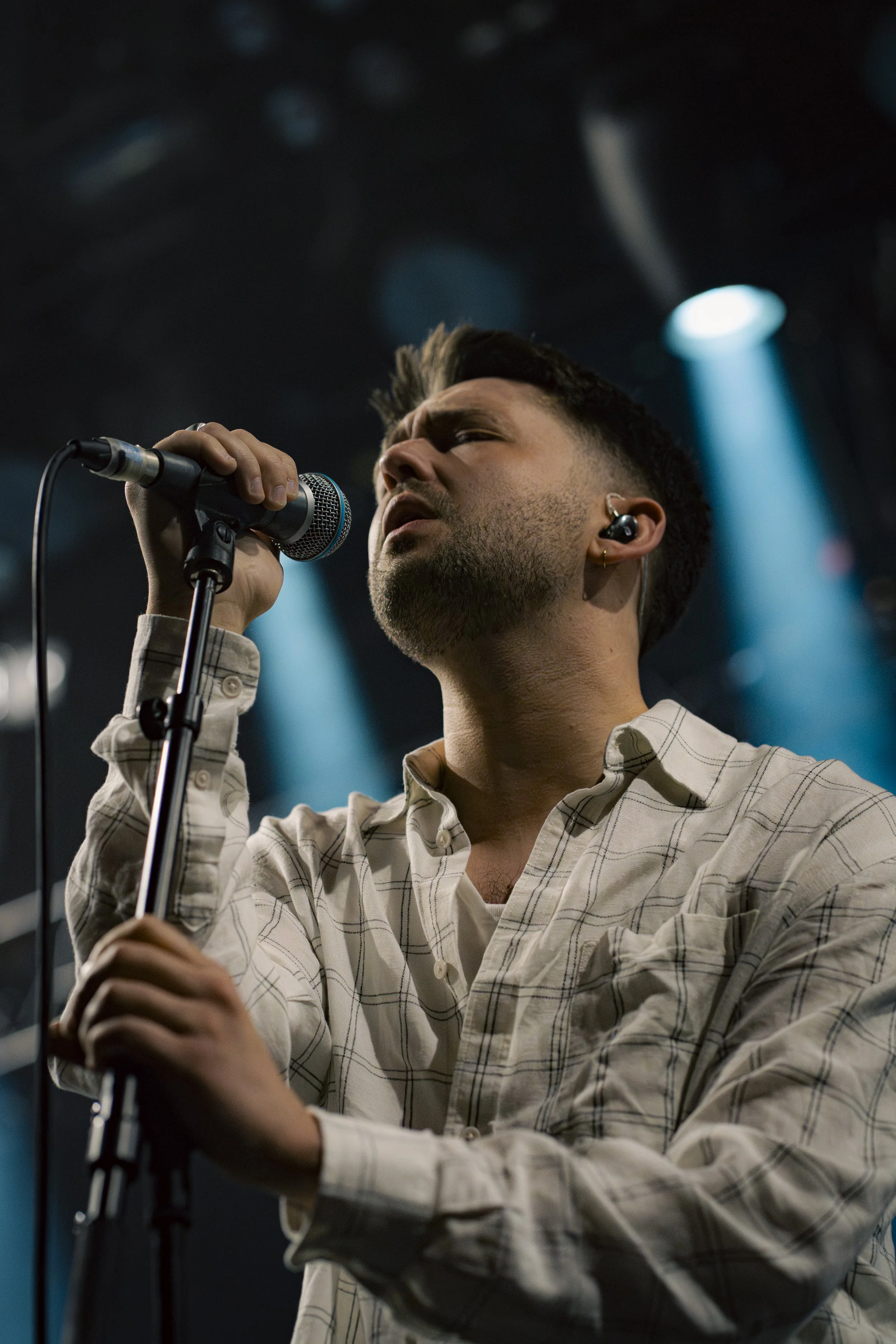 A man singing into a microphone on stage with stage lights shining behind him.