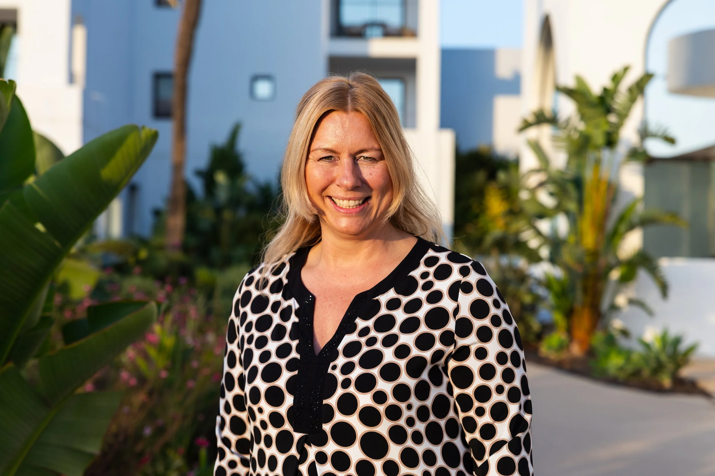 A woman with blonde hair smiling outdoors, wearing a black and white polka dot blouse, with modern apartment buildings and lush green plants in the background.