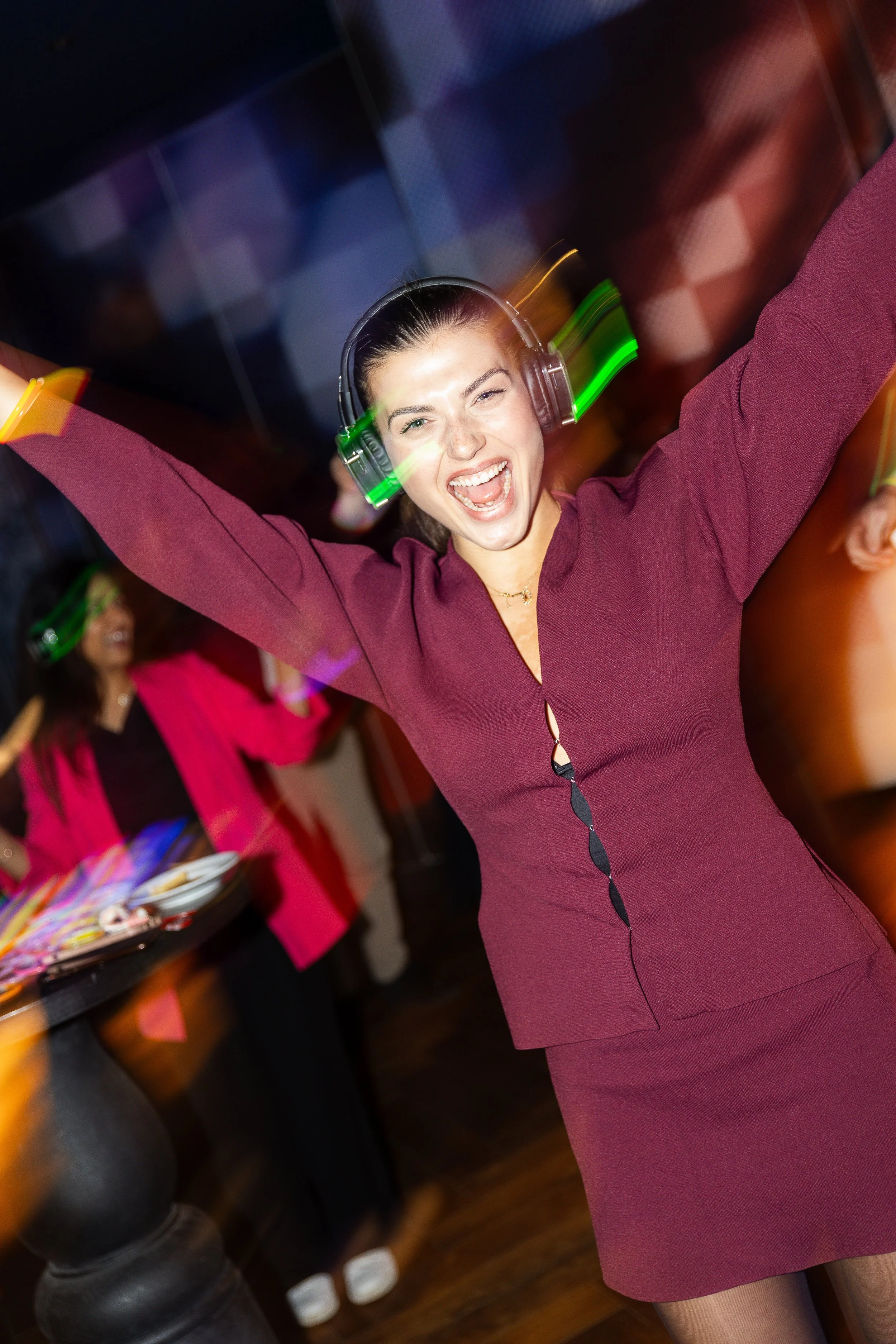 A woman in a burgundy dress with arms raised, wearing headphones, smiling and dancing at a lively party or celebration.