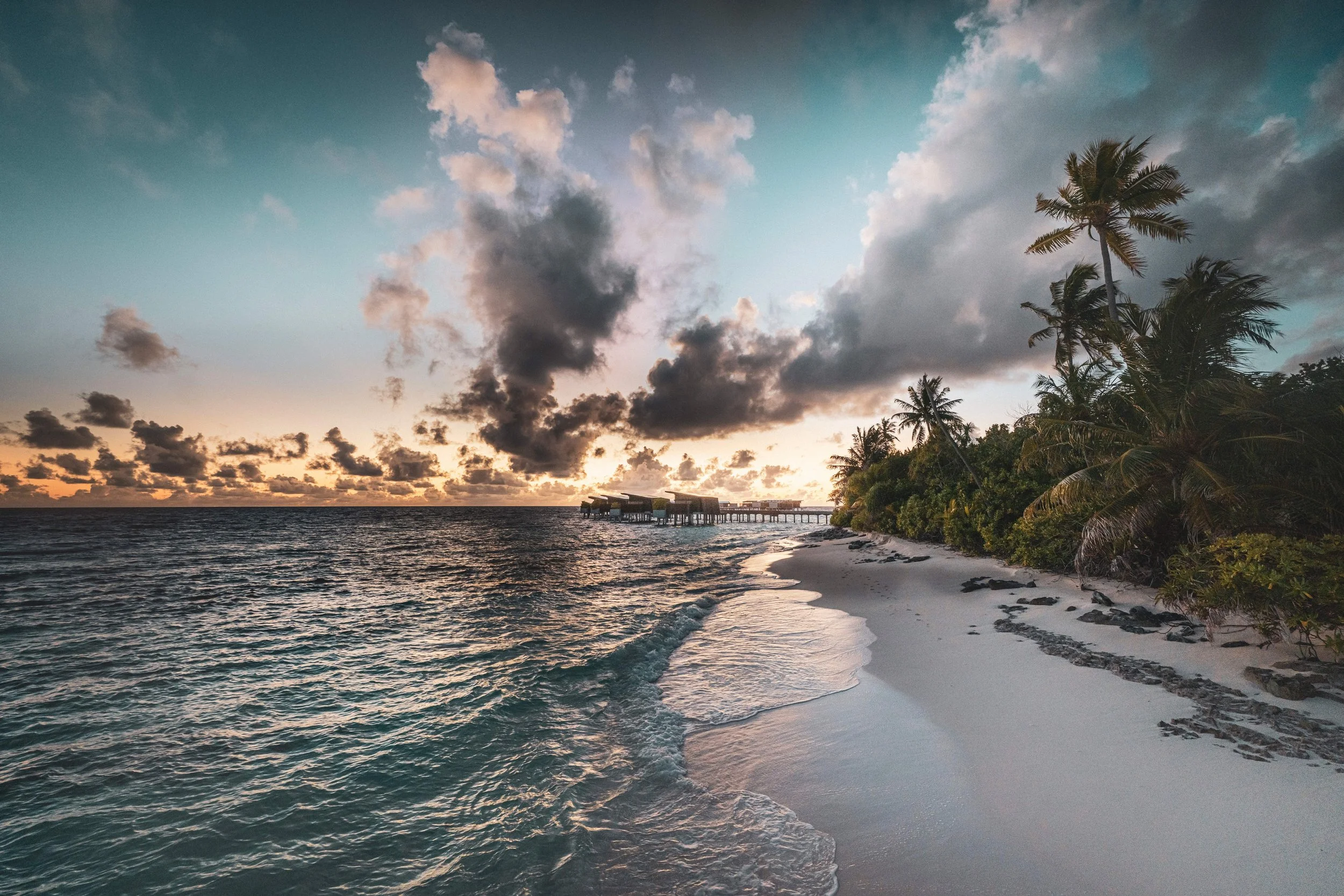 A tropical beach scene at sunset with palm trees, white sand, and an overwater structure in the distance. Clouds are in the sky, and the ocean waves gently reach the shore.