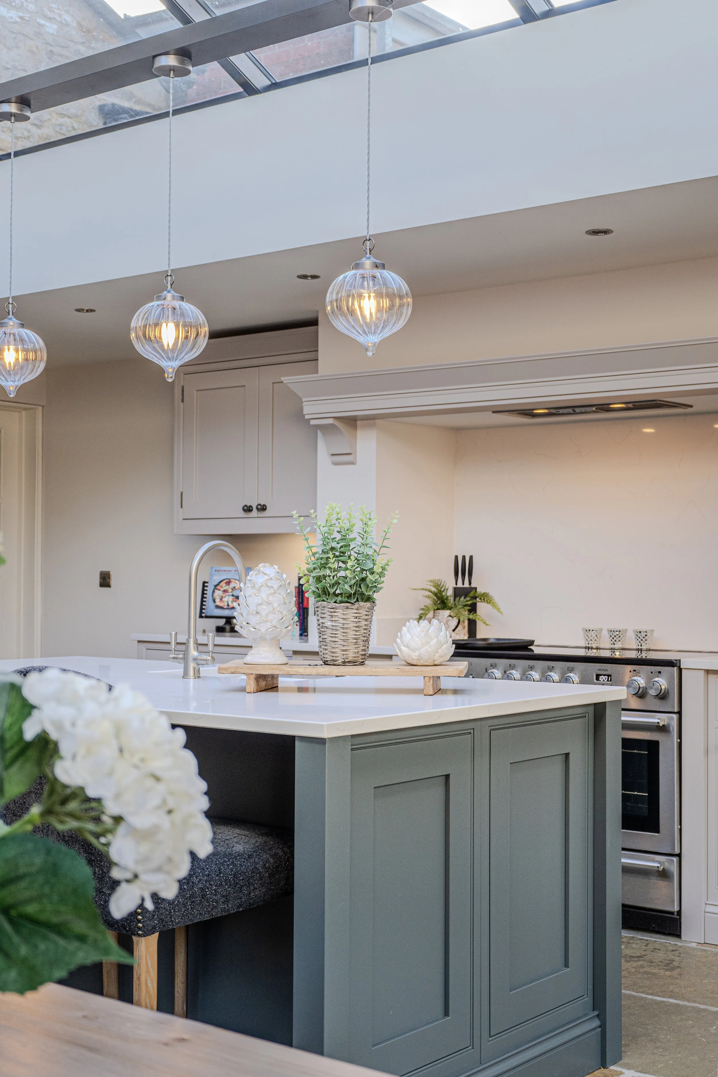 Modern kitchen with pendant lights, green island, white countertop, potted plants, and decorative objects.