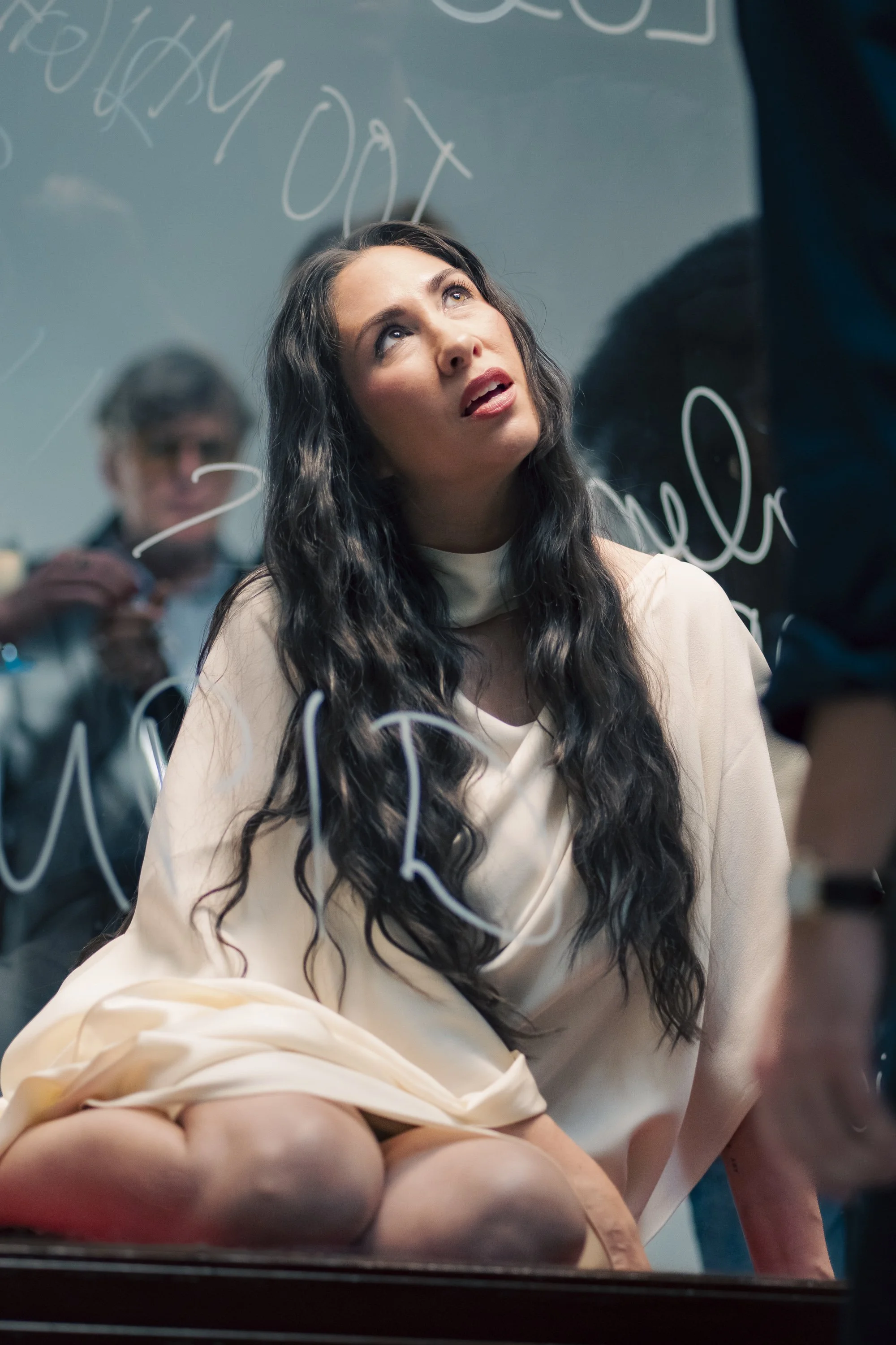 A woman with long wavy black hair, wearing a cream-colored dress, kneeling with her hands on the floor, looking up at a glass board with writing on it, with a person in the background taking notes.
