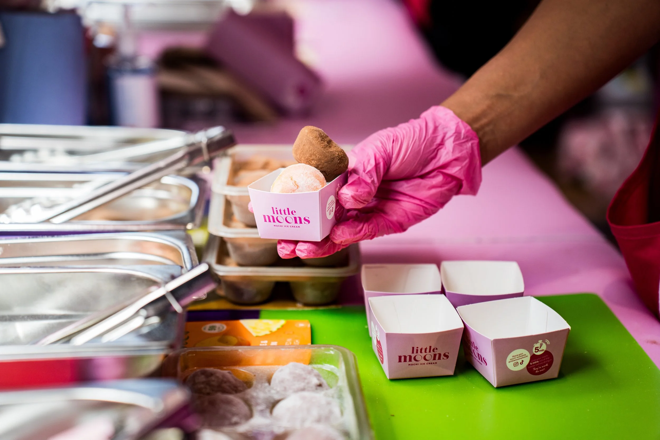 A person wearing pink gloves is holding a small paper cup of ice cream, with the label "little moons." The ice cream cups are on a colorful green and pink table, with other cups and containers of different ice cream flavors visible nearby.
