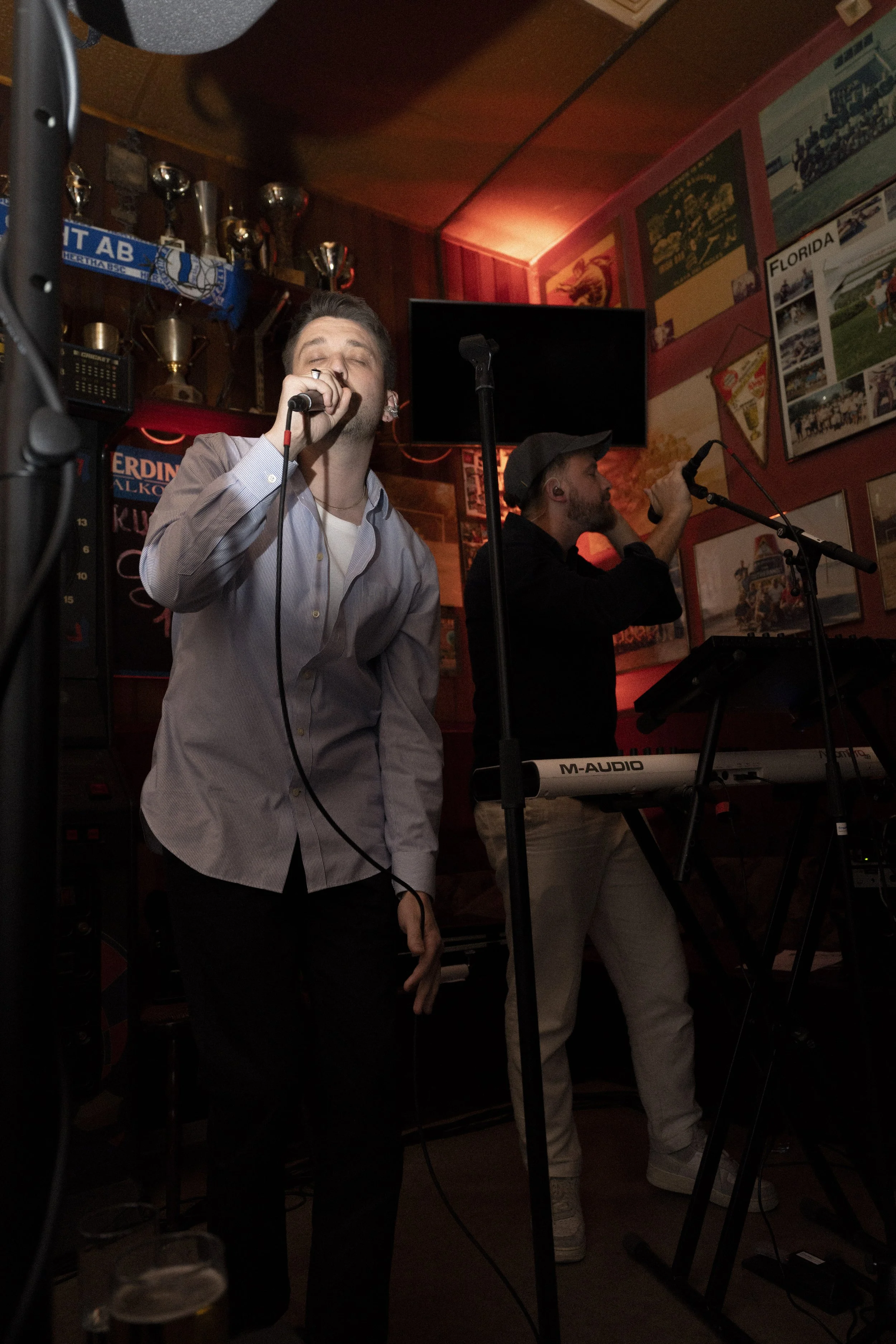 Two male musicians singing into microphones in a dimly lit bar or pub, with them standing near a keyboard, with a wall decorated with framed pictures and trophies in the background.
