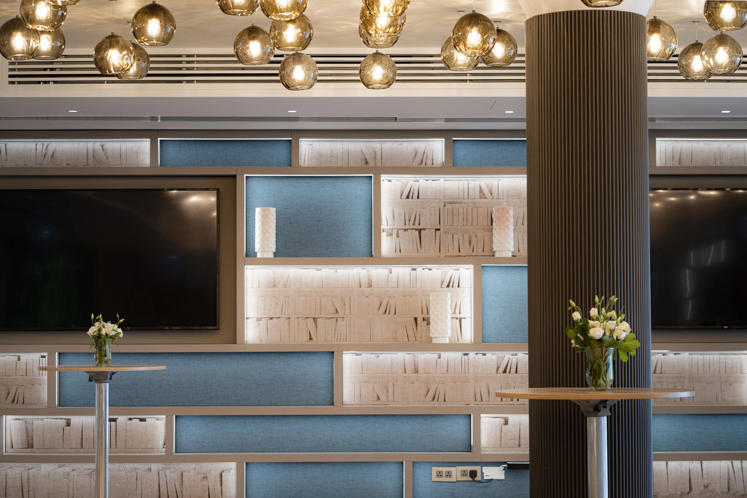 Interior of a modern lounge or hotel lobby with decorative lighting fixtures, blue padded panels, bookshelves, lamps, and flower arrangements on tables.