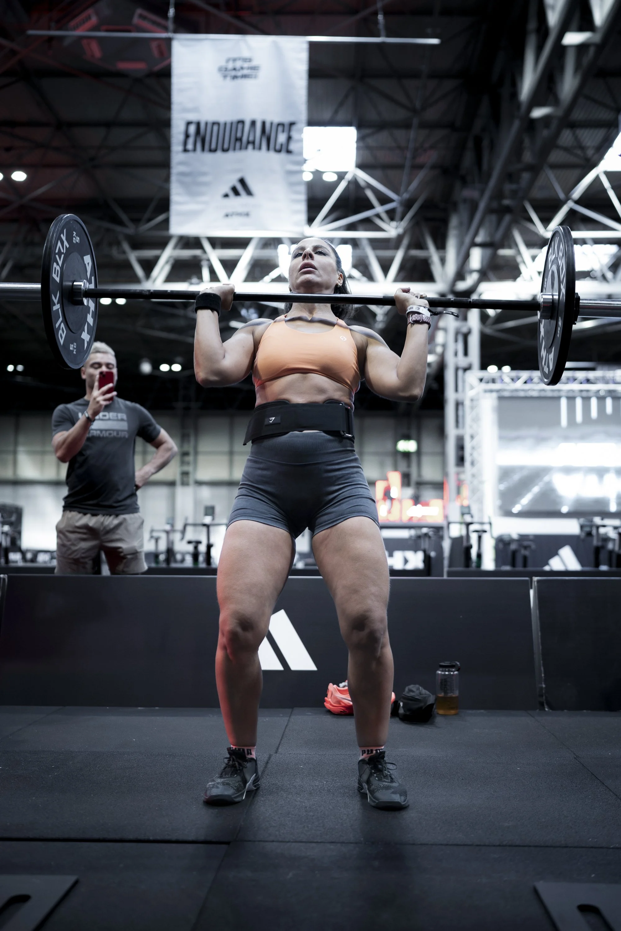 A woman weights lifting in a gym, holding a barbell with weights above her shoulders, with a man taking a photo in the background and a banner that says 'ENDURANCE' hanging from the ceiling.